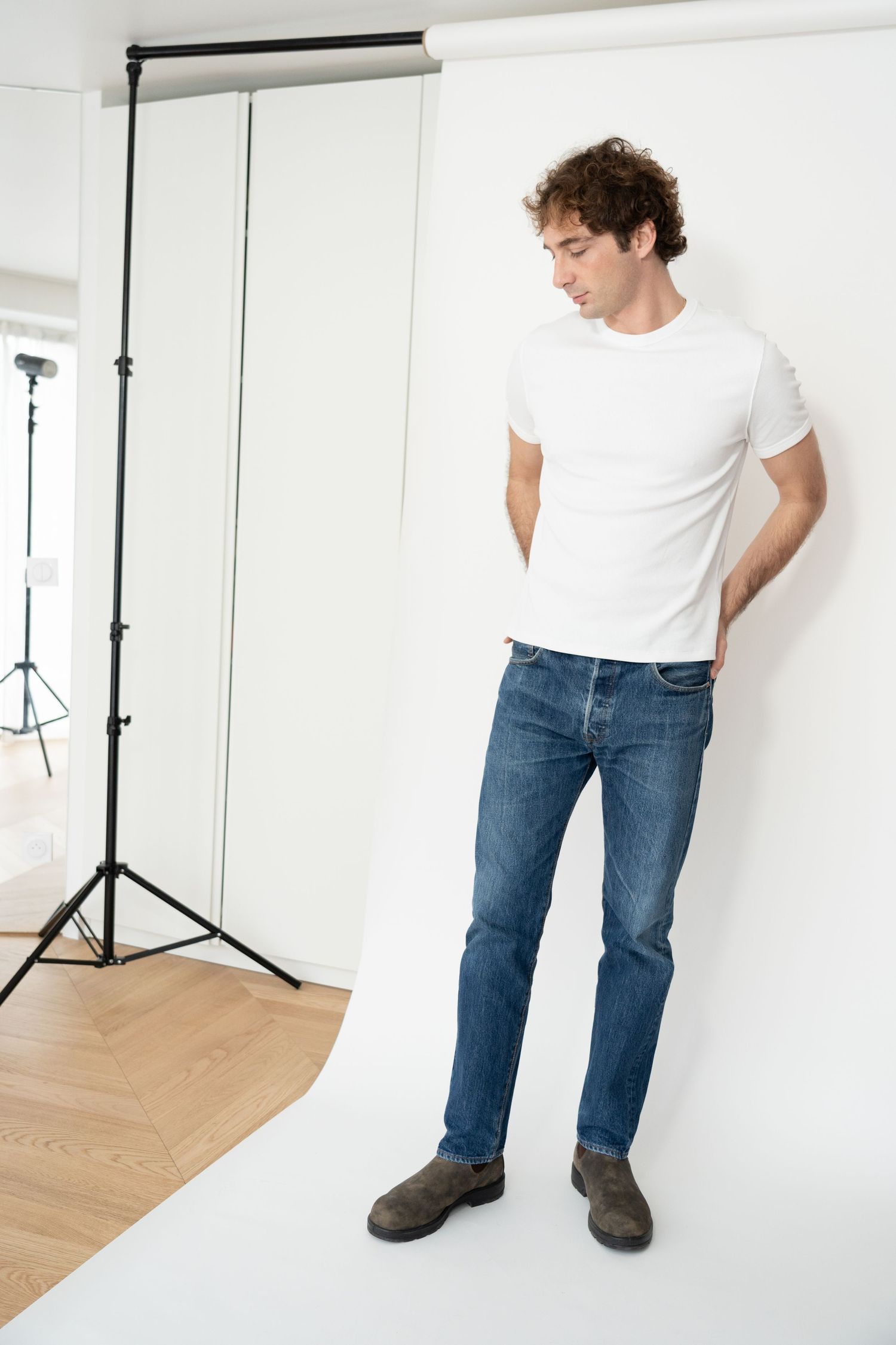 Man in white t-shirt and blue jeans posing in a photography studio with white backdrop and lighting equipment.