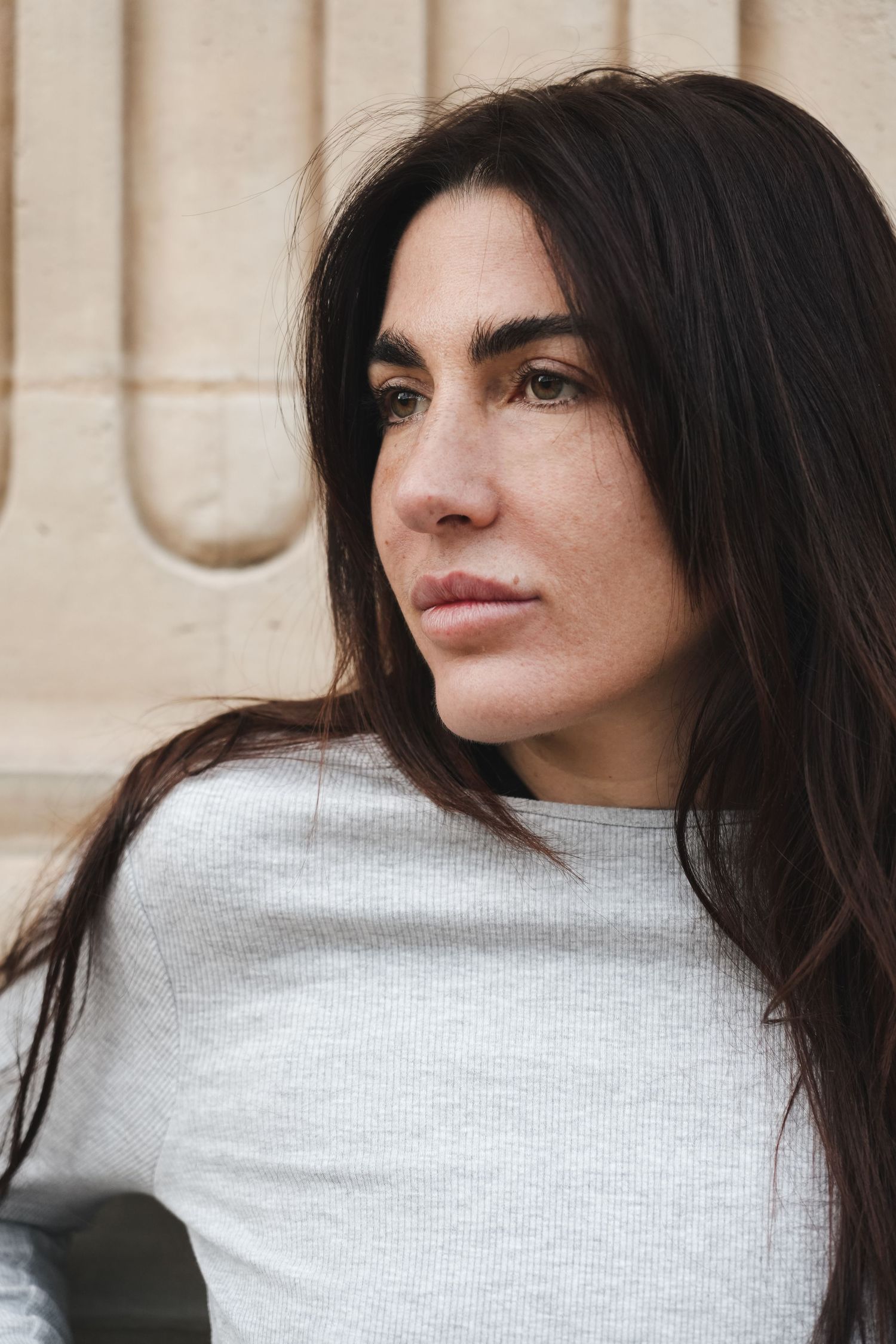 A dark-haired woman in a white top gazes thoughtfully to the side against a stone wall backdrop.