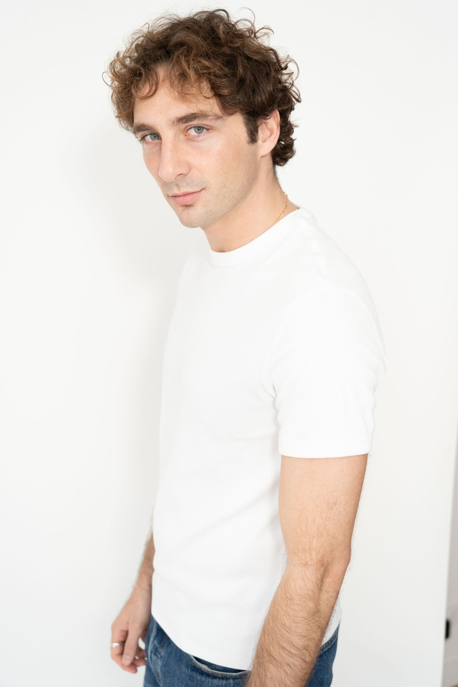 Young man with curly hair wearing white fitted t-shirt and jeans in side profile pose.