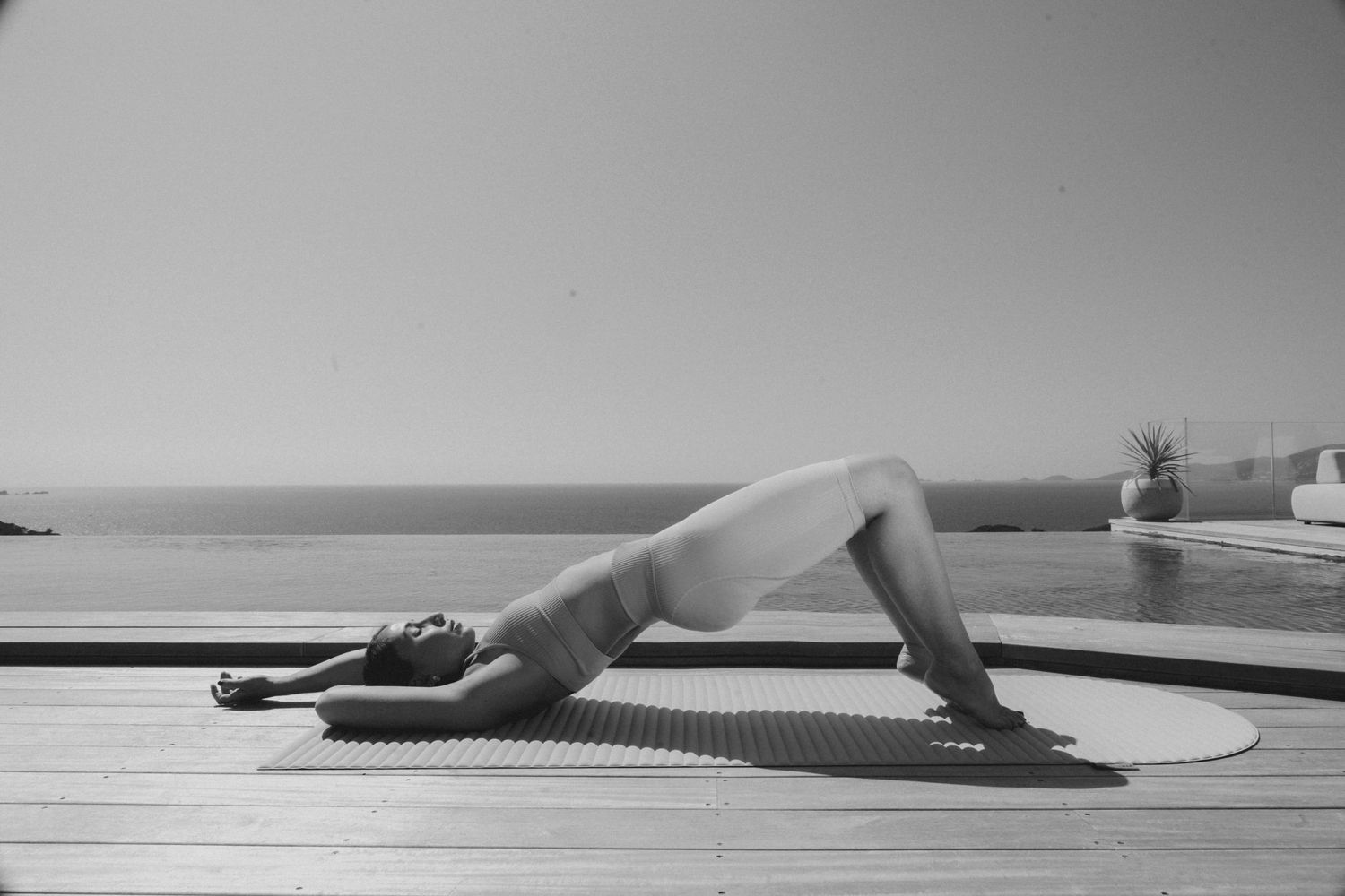 Person practicing yoga in a reverse plank pose on a mat by an infinity pool overlooking the ocean.