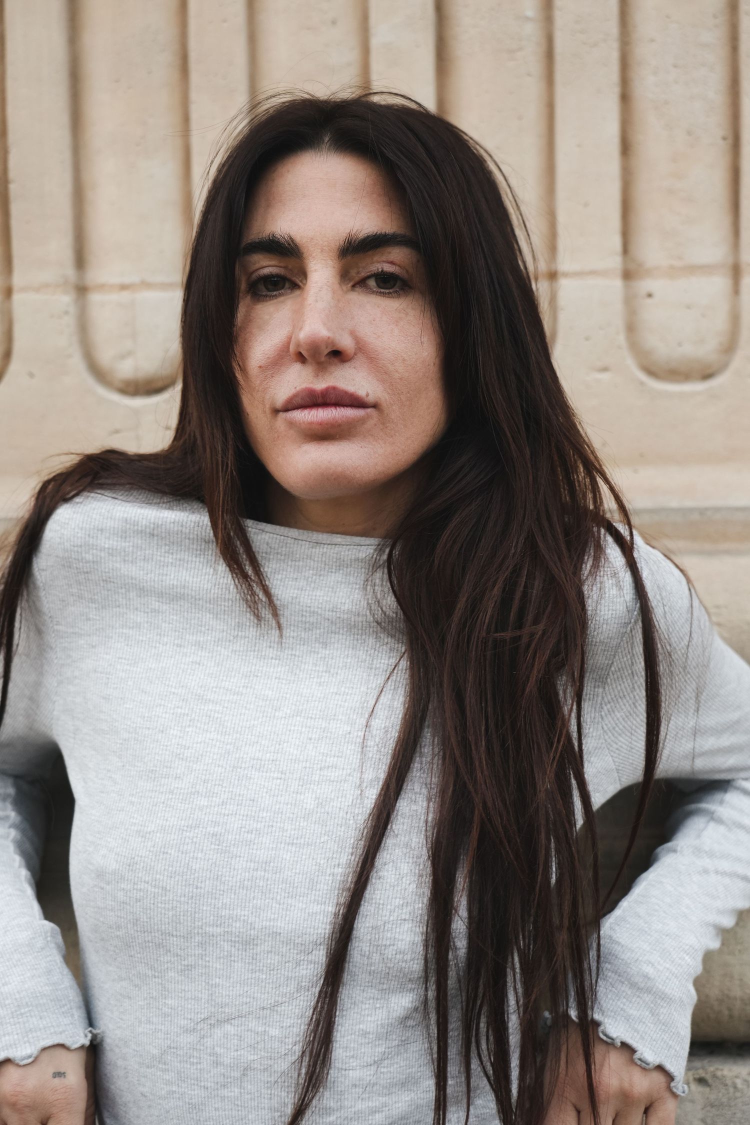Dark-haired woman in gray sweatshirt looking directly at camera with serious expression against stone wall.