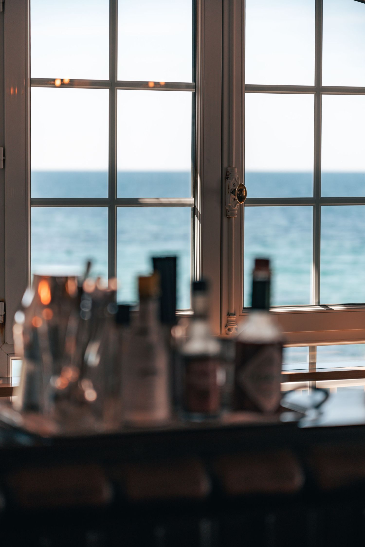 Blurred bottles on a bar counter with a white-framed window overlooking a serene ocean view in the background.