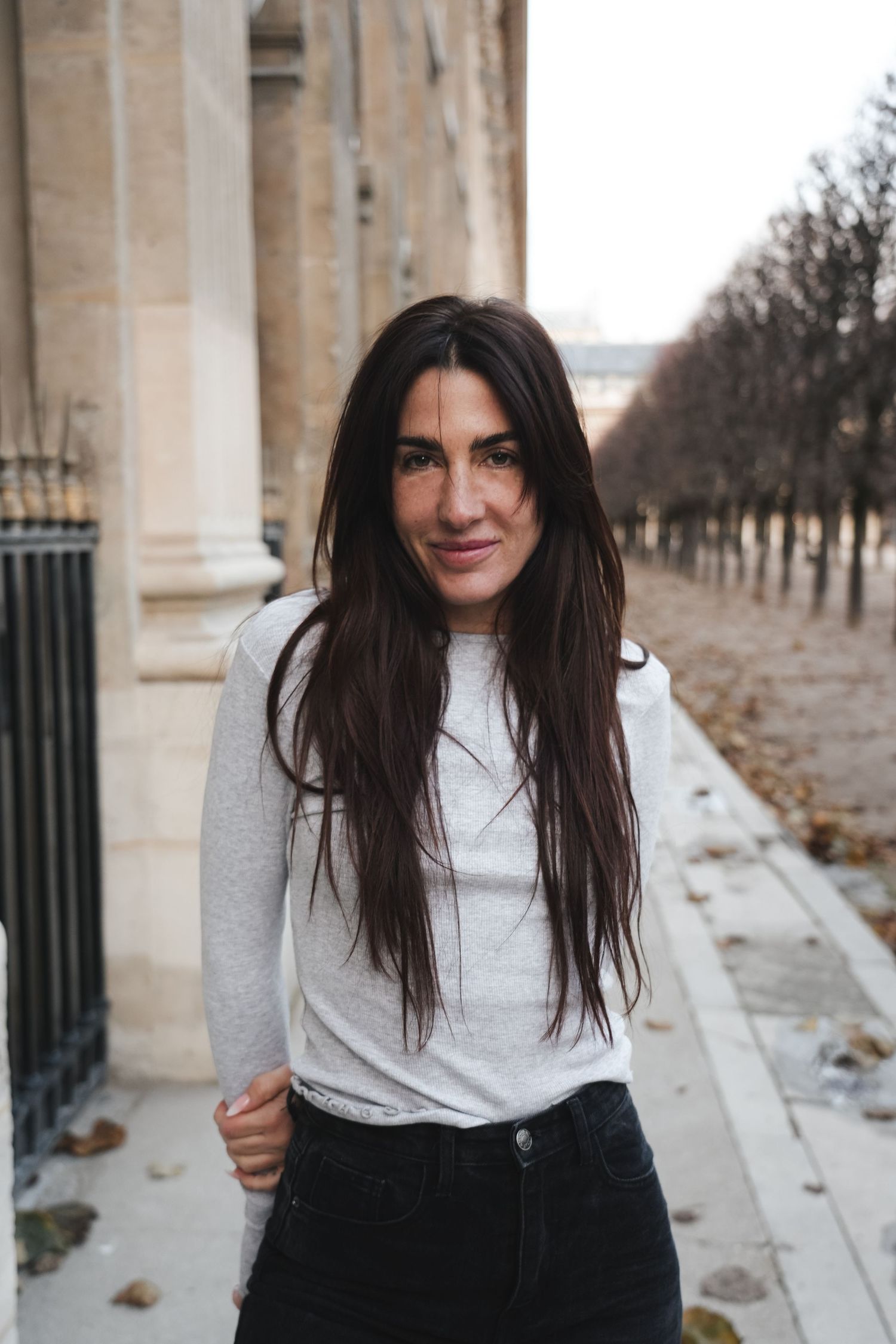 Young woman with long dark hair in gray top and black jeans smiling on a Parisian sidewalk in winter.