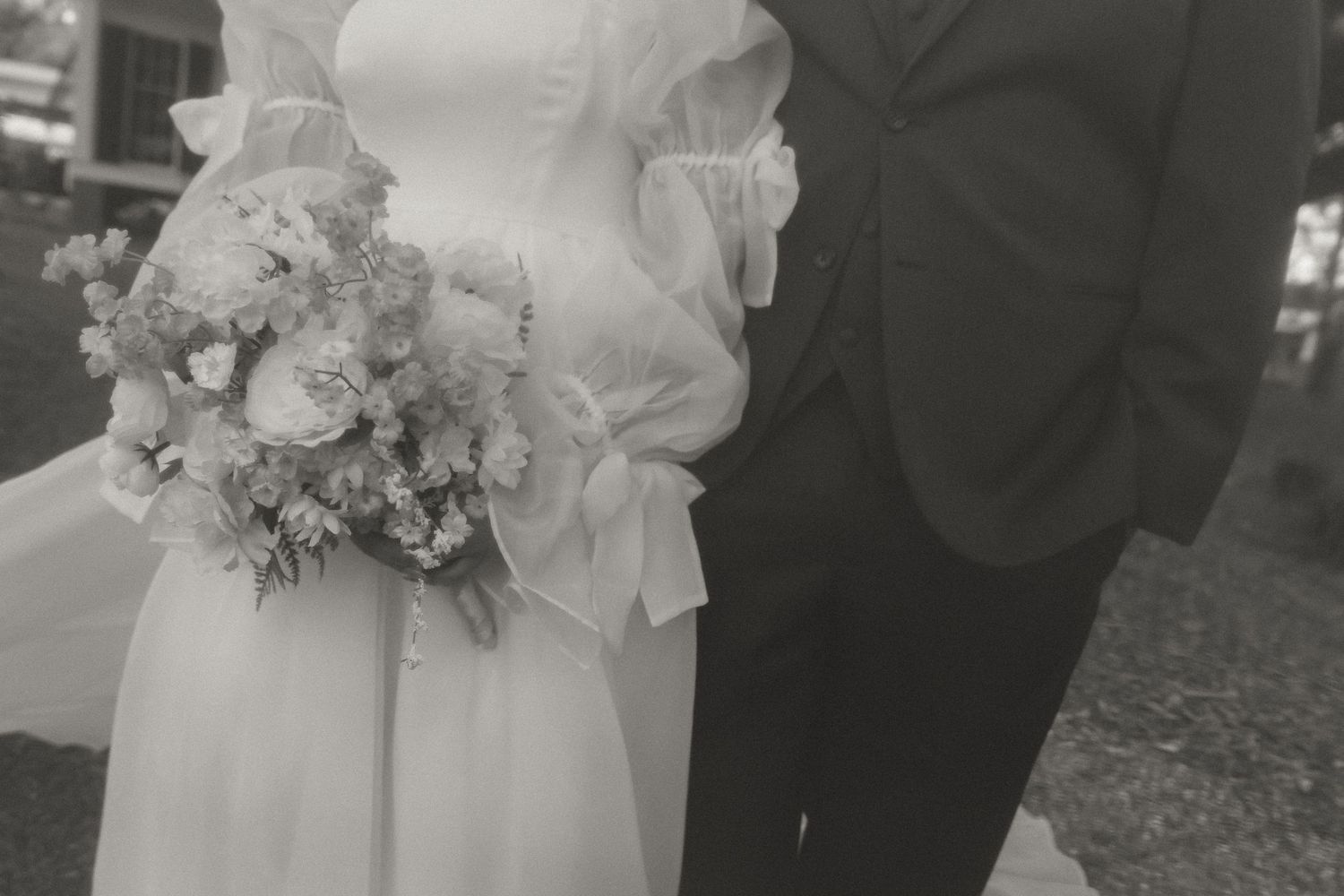 Black and white close-up of bride holding lush floral bouquet with groom in dark suit beside her at wedding.