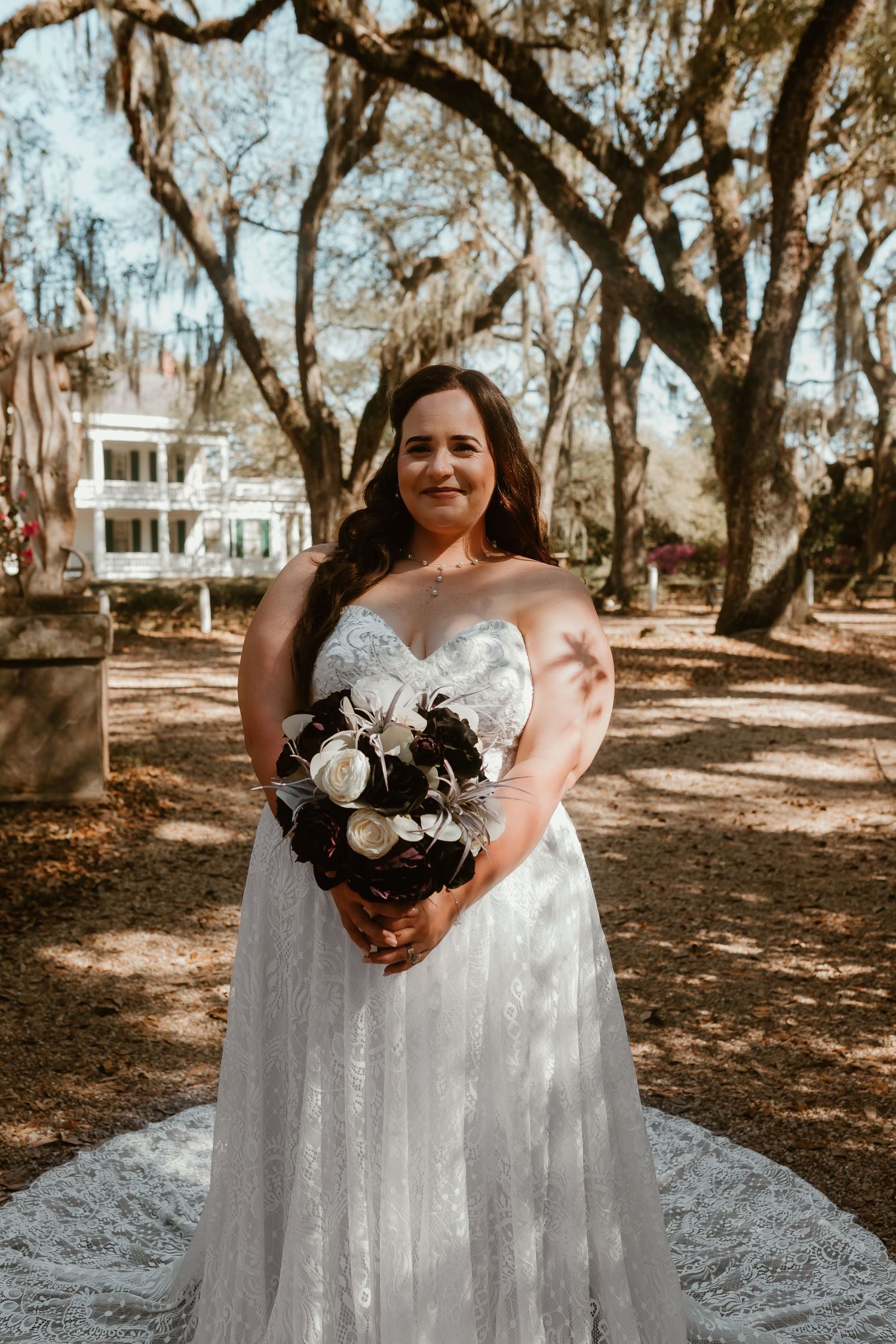Smiling bride in strapless lace wedding gown holding bouquet under Spanish moss-draped oak trees.