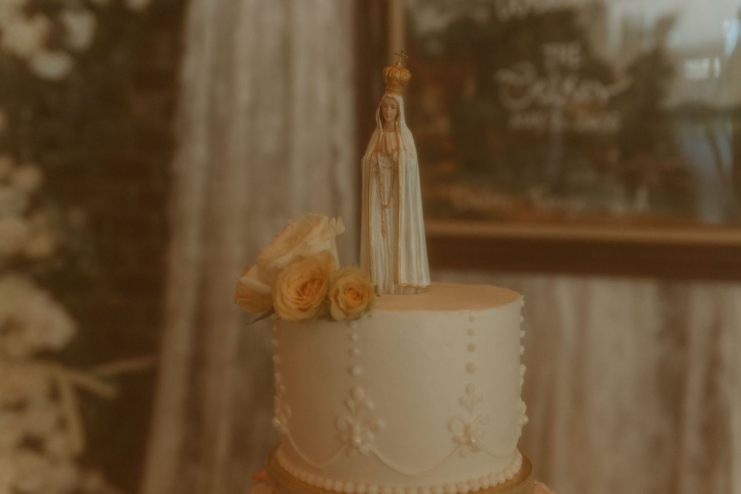 Elegant white wedding cake topped with a Our Lady of Fatima figurine and yellow roses.