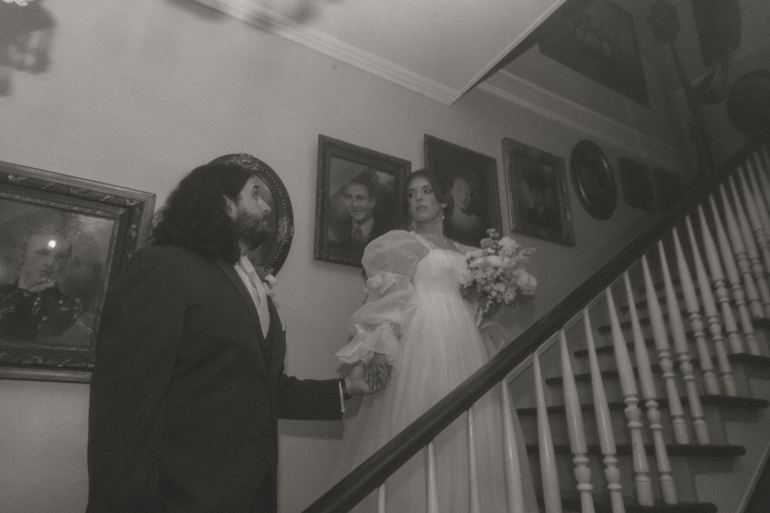 Black and white wedding photo of bride on staircase holding bouquet with groom standing below in elegant home.