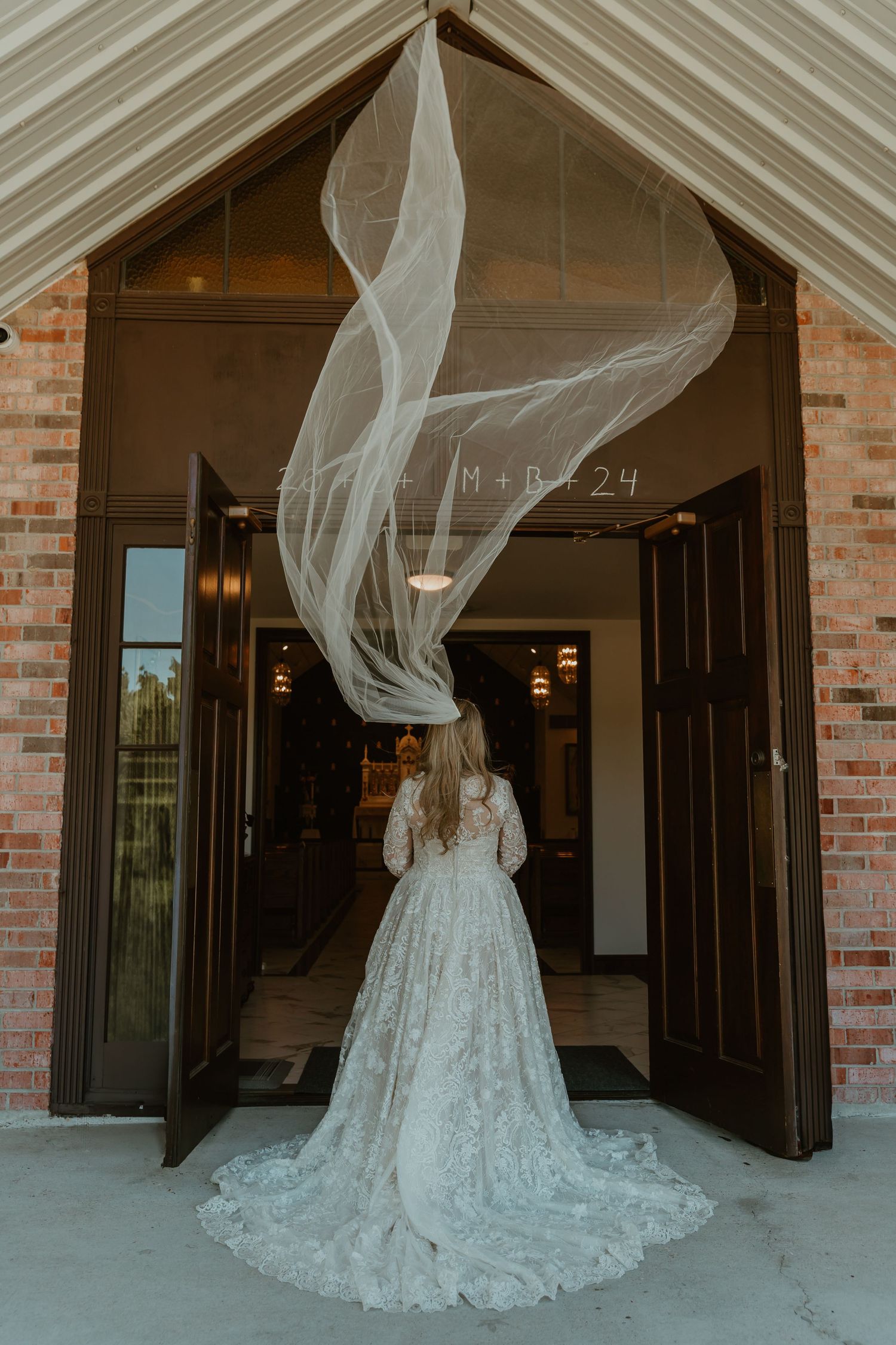 Bride in lace gown with veil flowing dramatically at church doors.