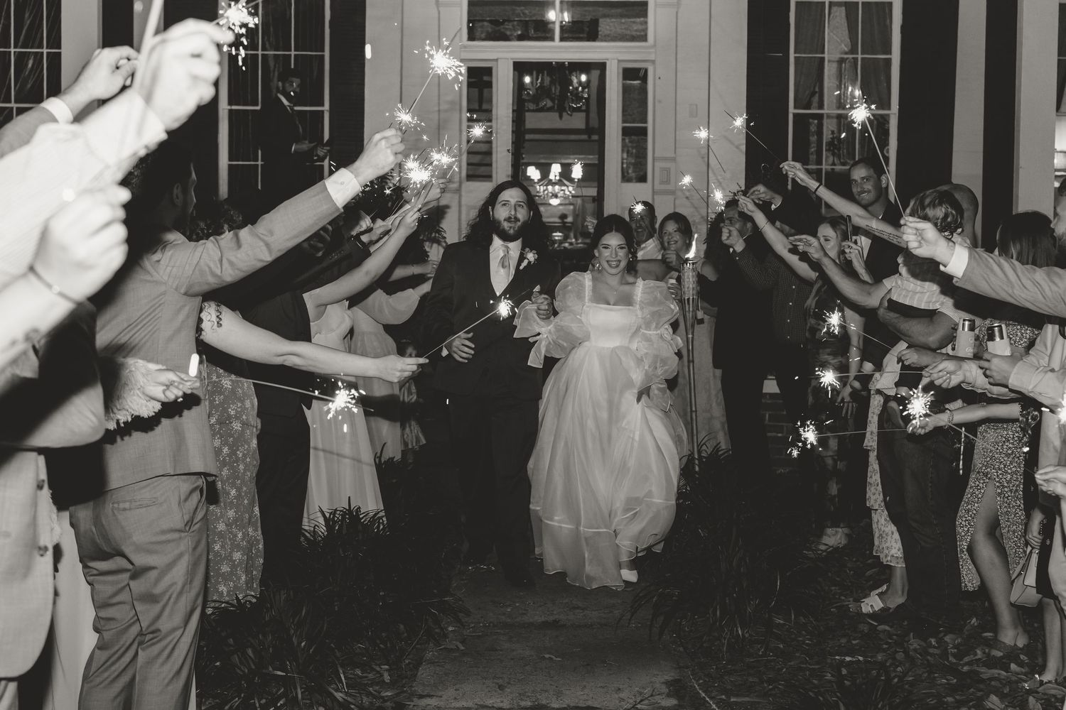 Black and white photo of newlyweds walking through a sparkler tunnel surrounded by celebrating guests.