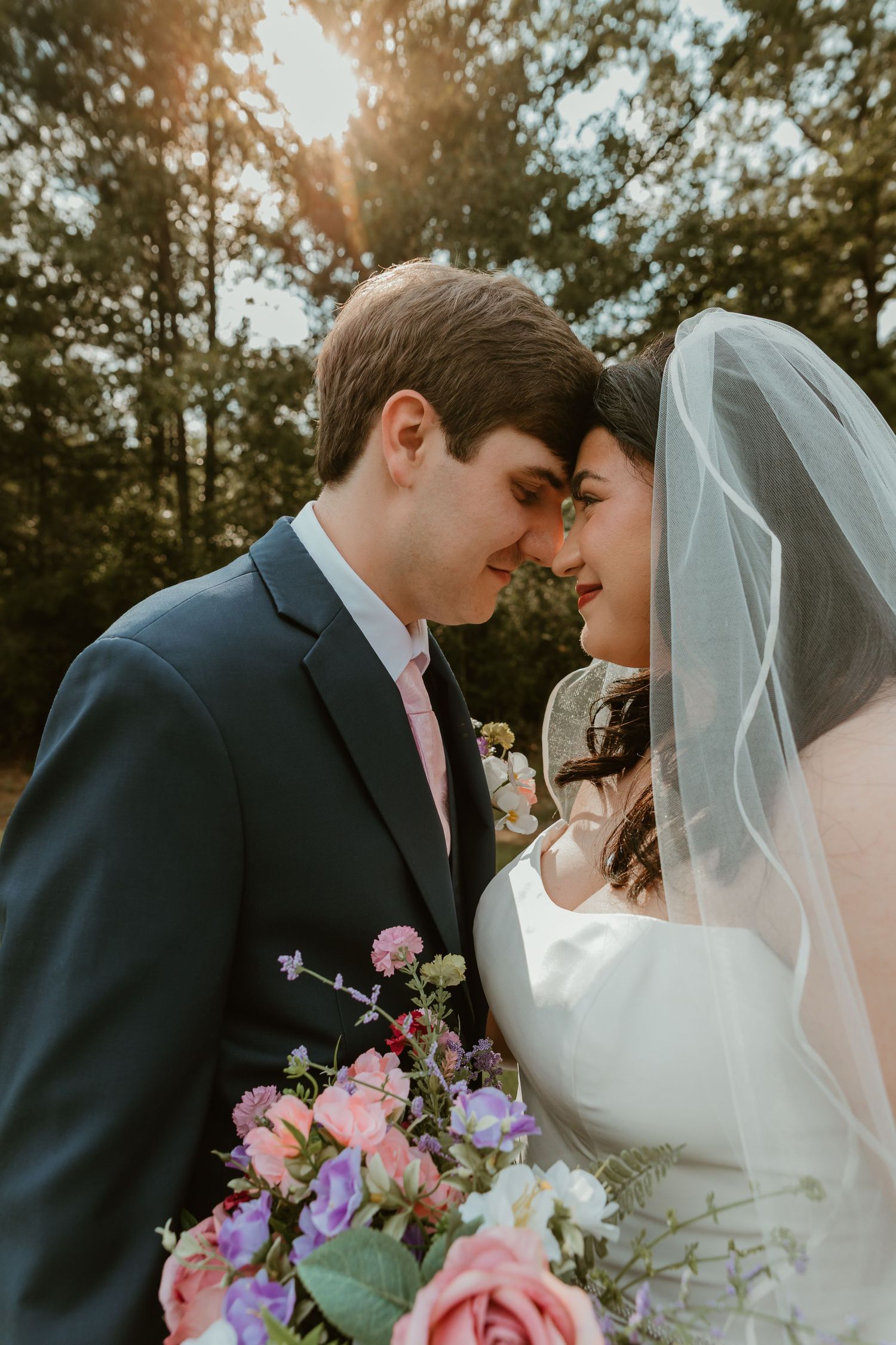 Bride and groom touch foreheads romantically in golden sunset light with floral bouquet in foreground.