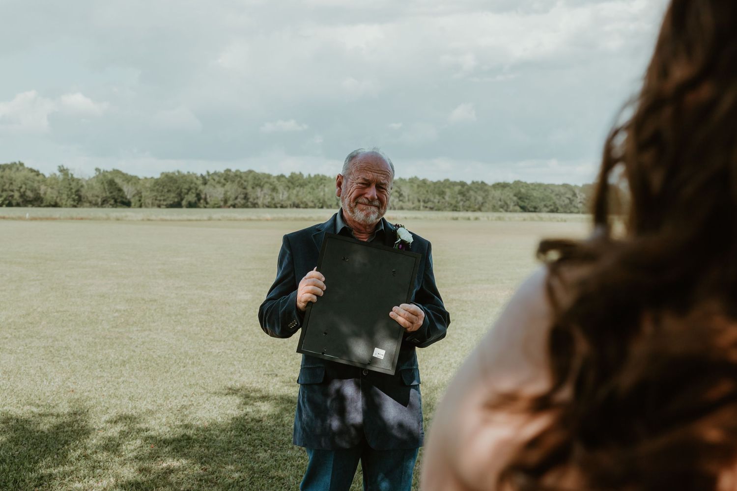 Smiling dad holds a young portrait of bride at an outdoor wedding ceremony in a grassy field.