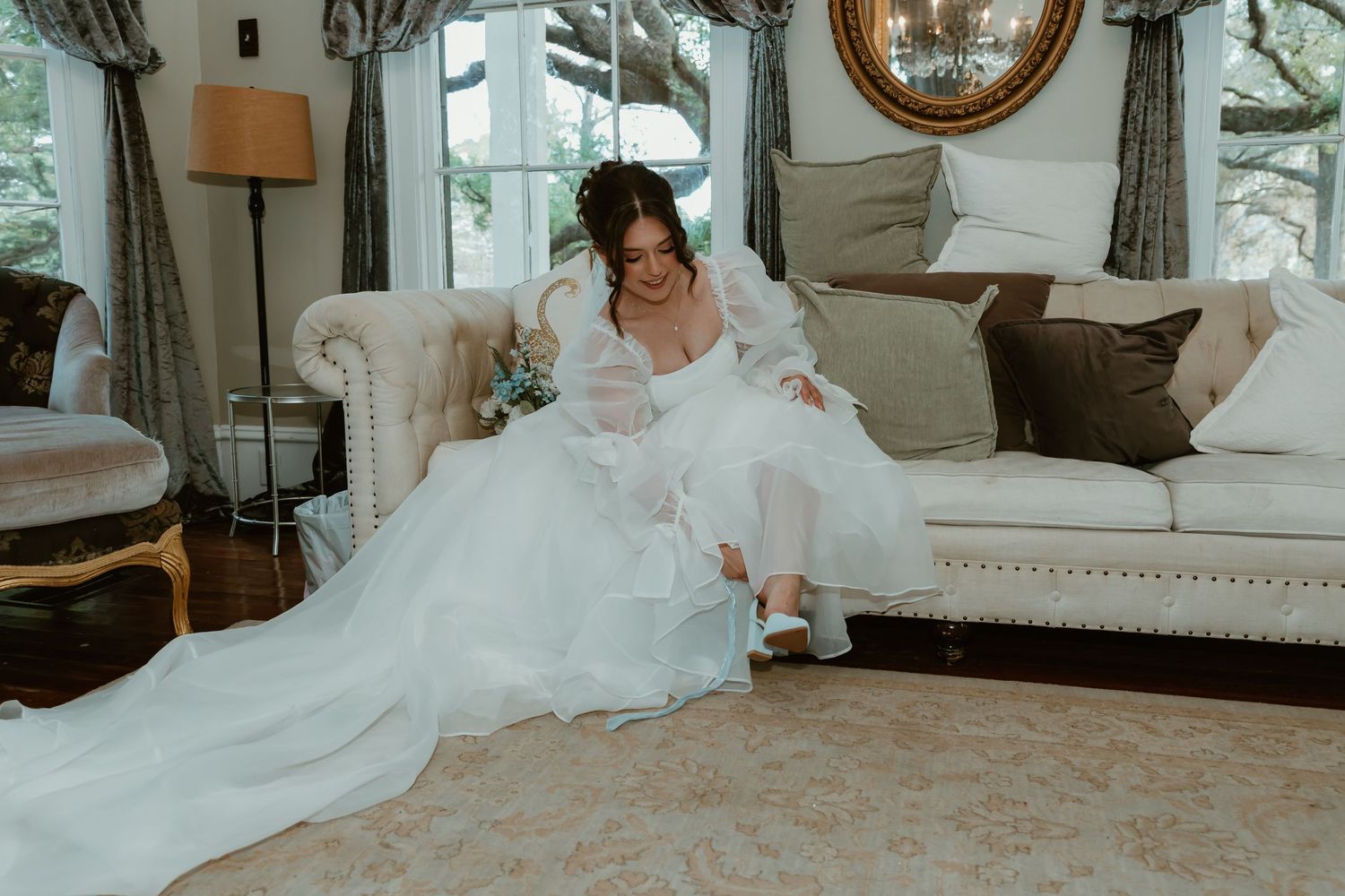Bride in flowing white wedding gown sitting on cream sofa putting on light blue bridal shoes.