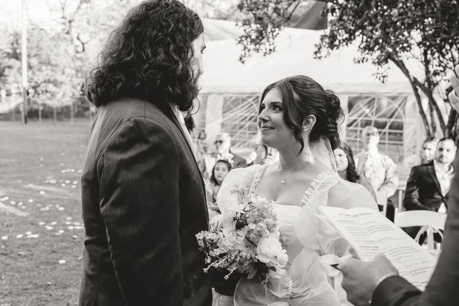 Black and white photo of a smiling bride holding a bouquet facing her long-haired groom at an outdoor wedding.