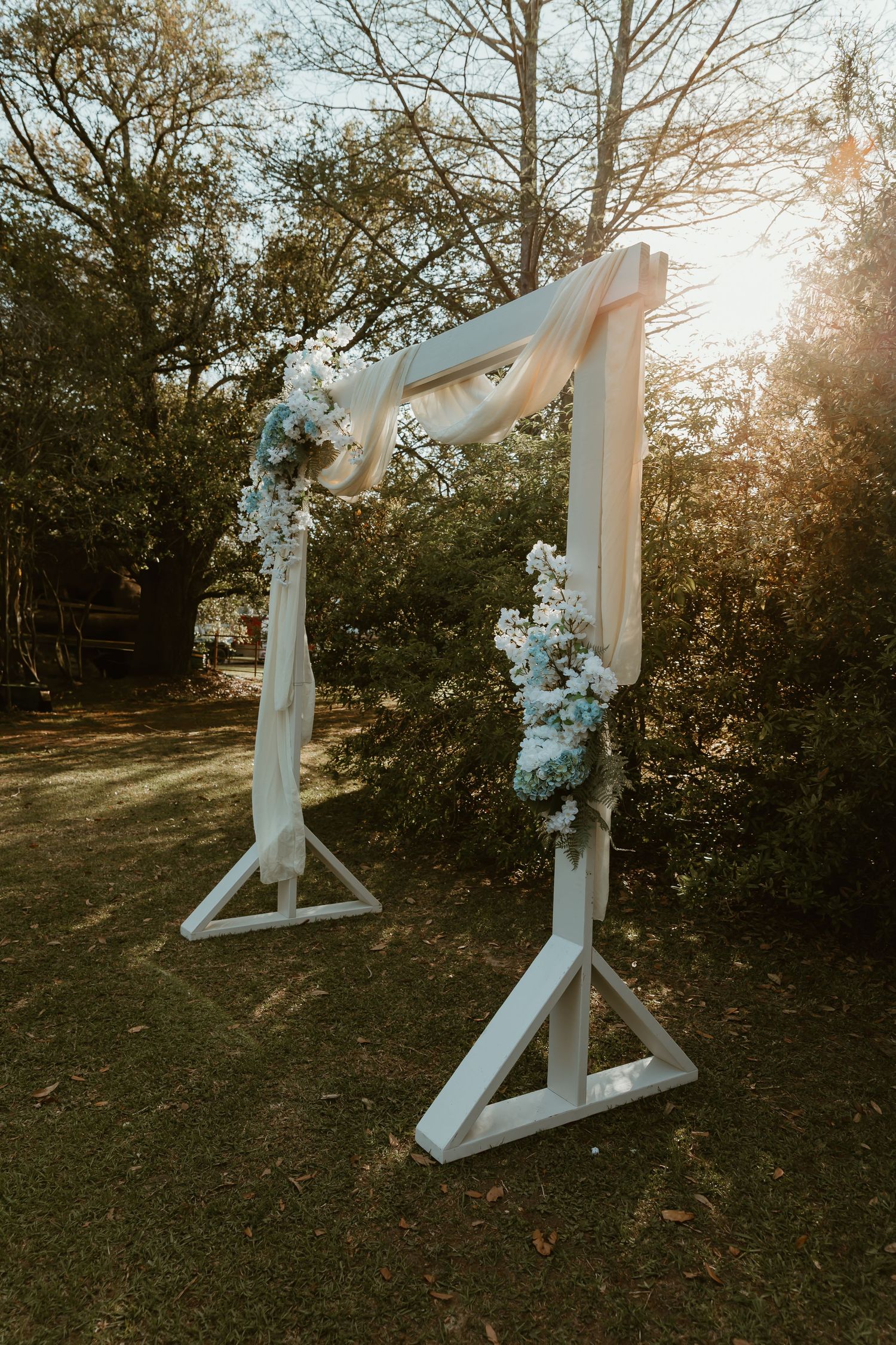 White wooden wedding arch adorned with blue and white floral arrangements glowing in golden sunlight.