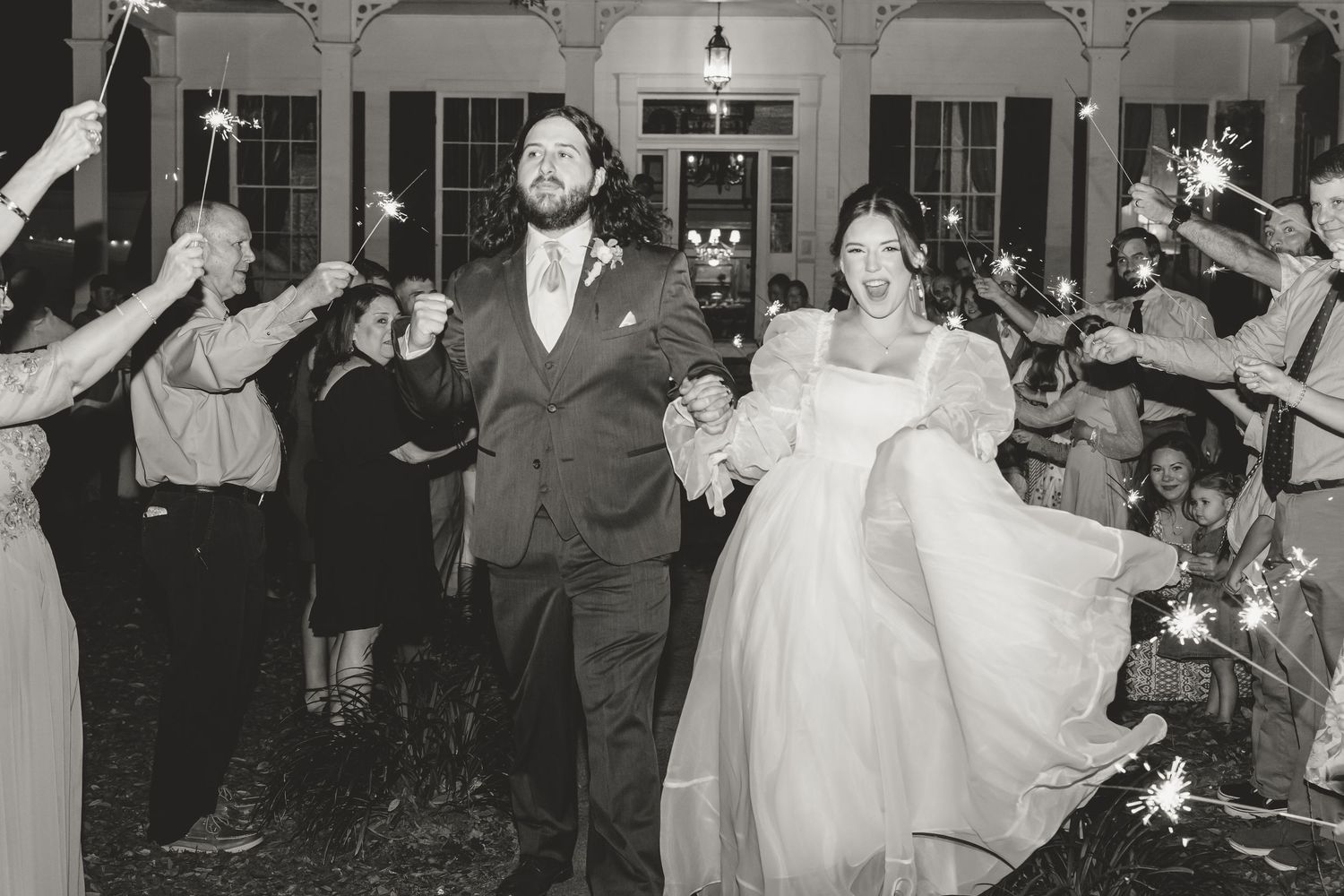 Black and white photo of newlyweds walking through sparkler exit with smiling wedding guests.