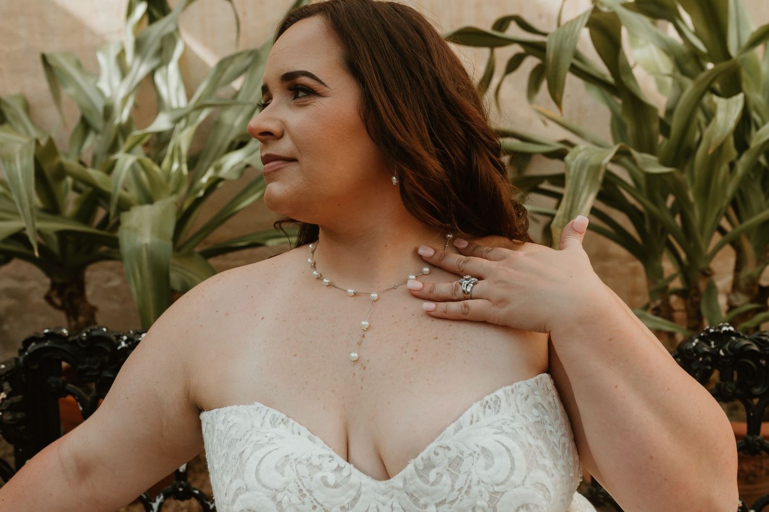 Bride in strapless lace wedding gown wearing pearl necklace and rings, posing among tropical plants.