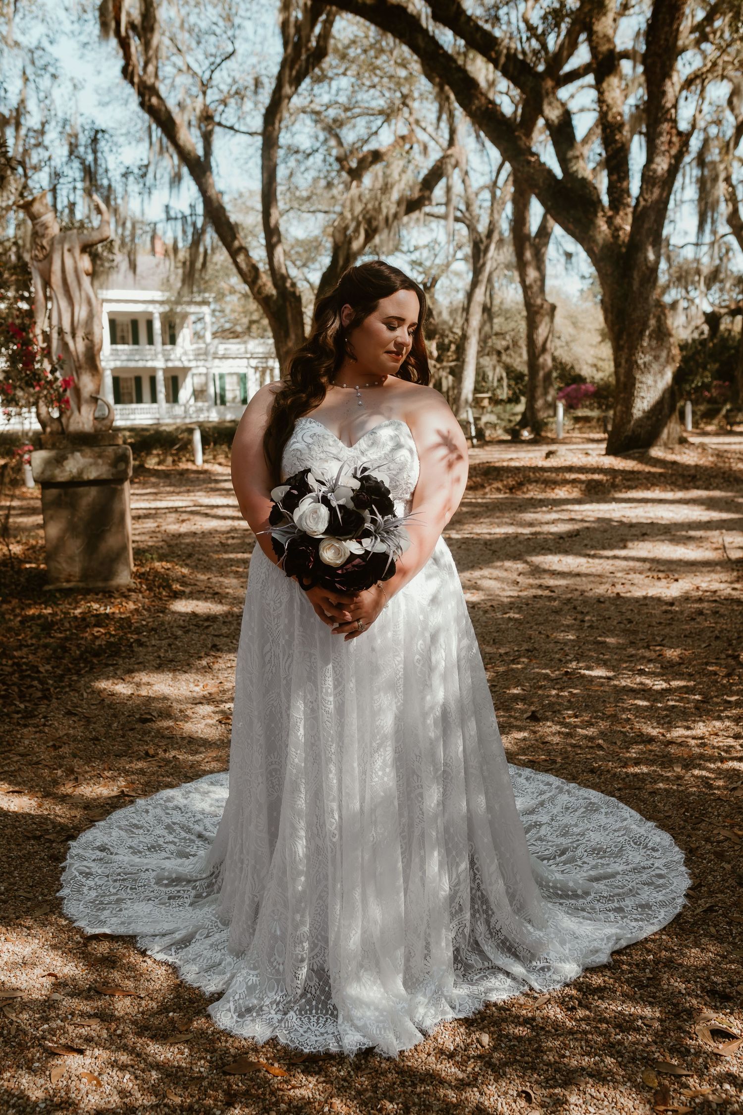 Bride in flowing lace strapless gown with cathedral train holding bouquet under moss-covered oak trees.