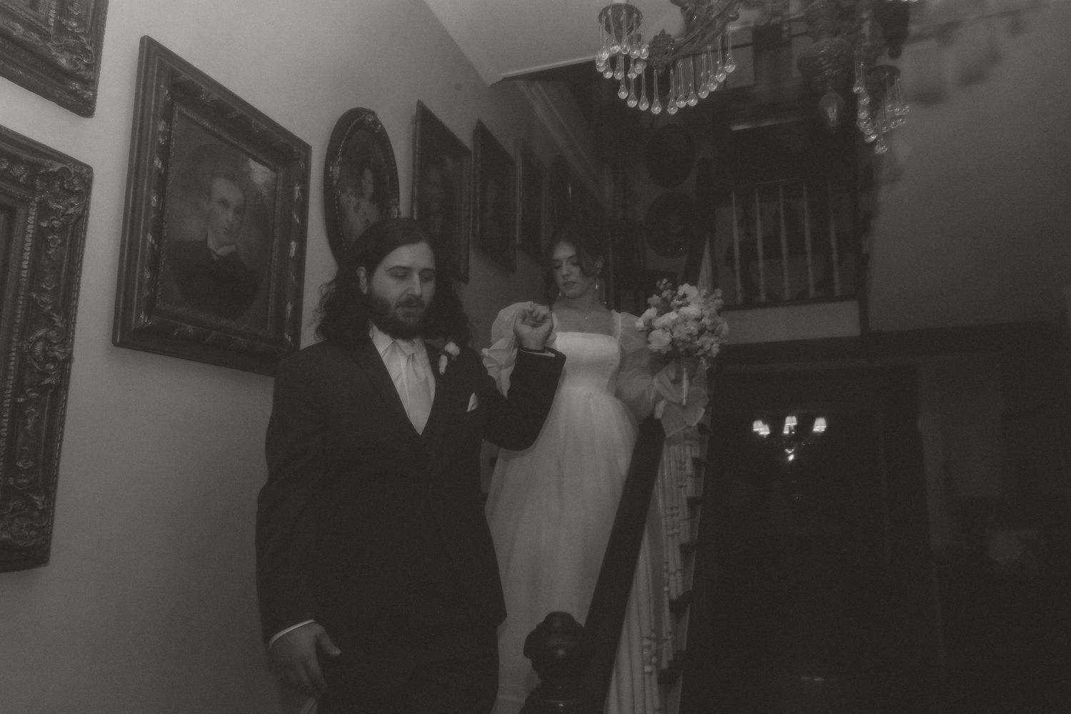 Black and white photo of groom leading bride down grand staircase adorned with portrait paintings at wedding venue.