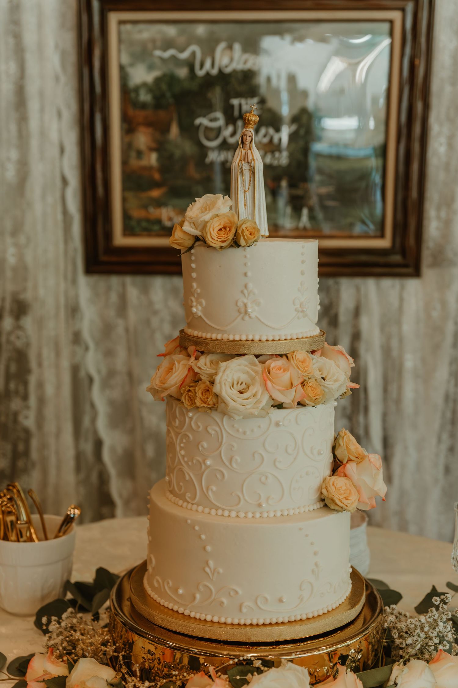 Elegant three-tier white wedding cake with roses and Virgin Mary topper.