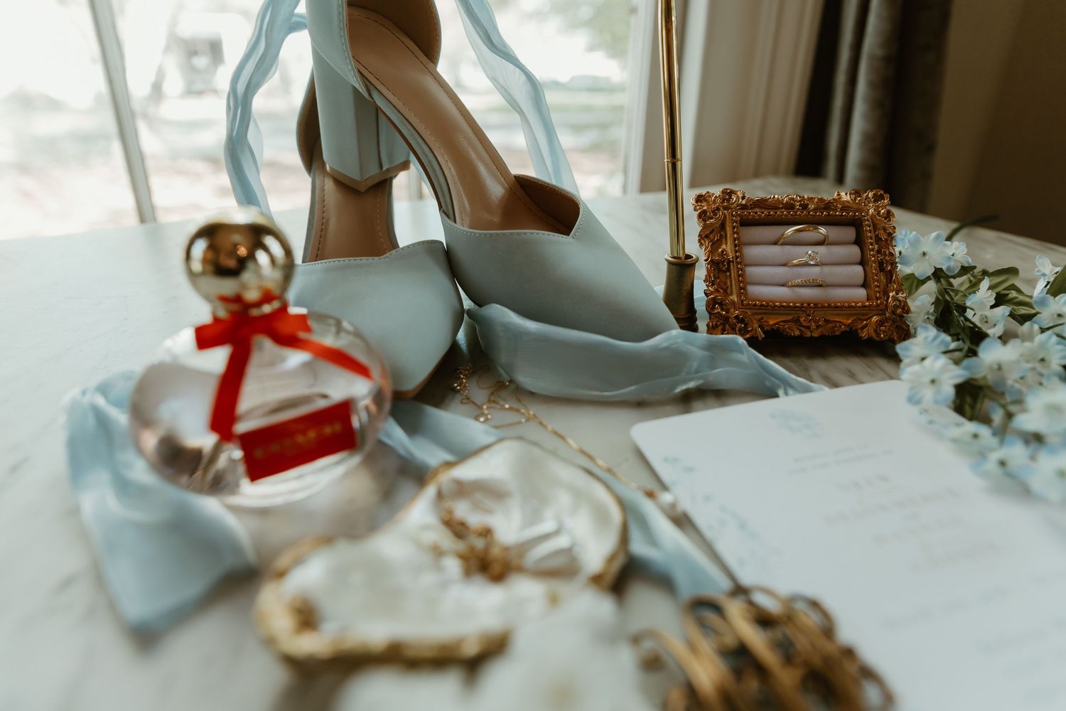 Wedding detail flat lay featuring blue block heels, perfume, rings in gold frame, and stationery.