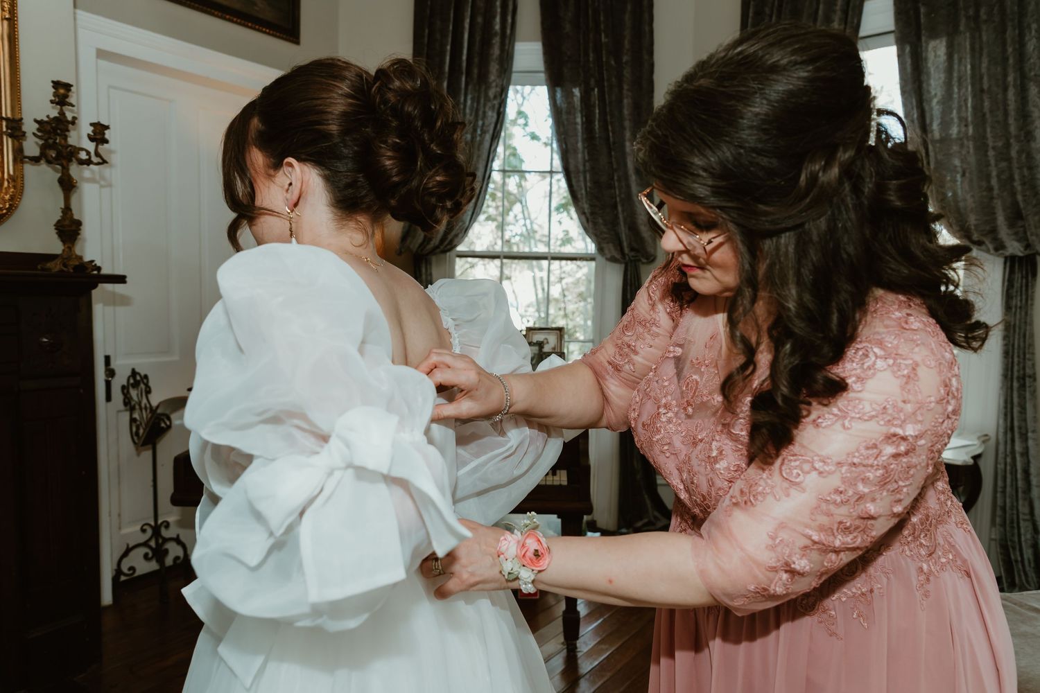 Mom fastening the back of a brides white wedding dress with a large bow.