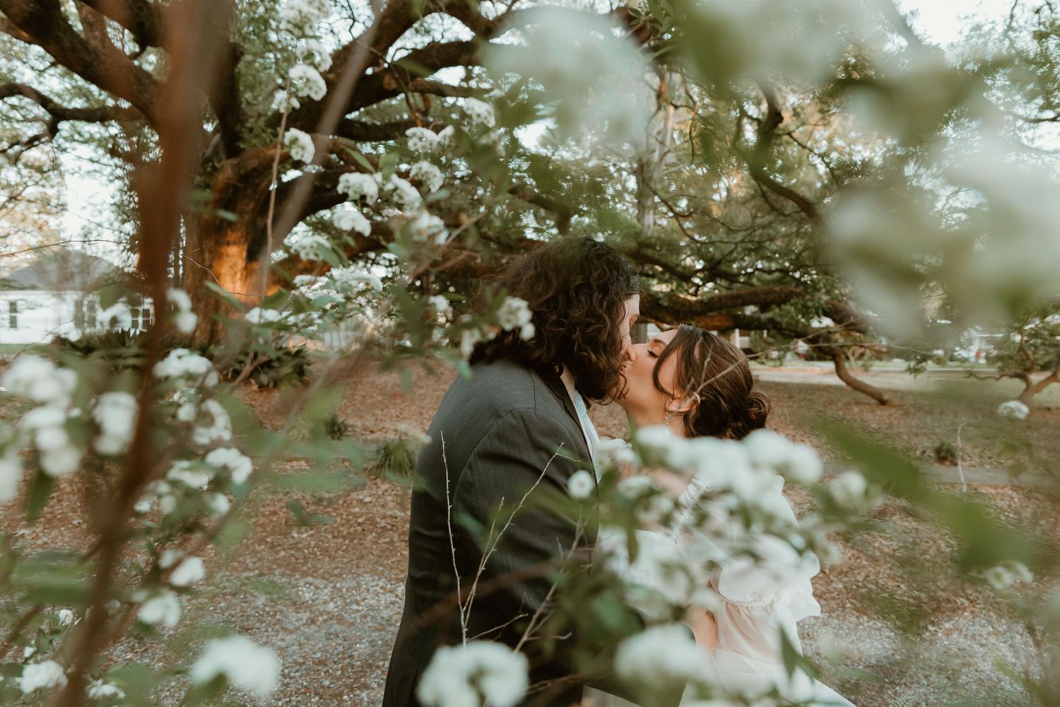 Couple shares a tender kiss surrounded by delicate white blossoms in garden setting.