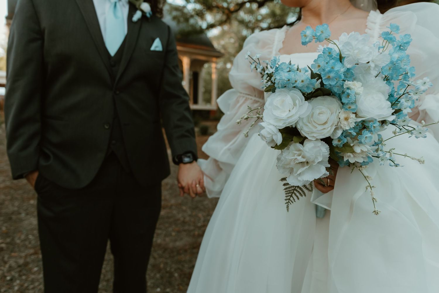 A bride and groom hold hands outdoors, bride carries a stunning bouquet of white and blue flowers.