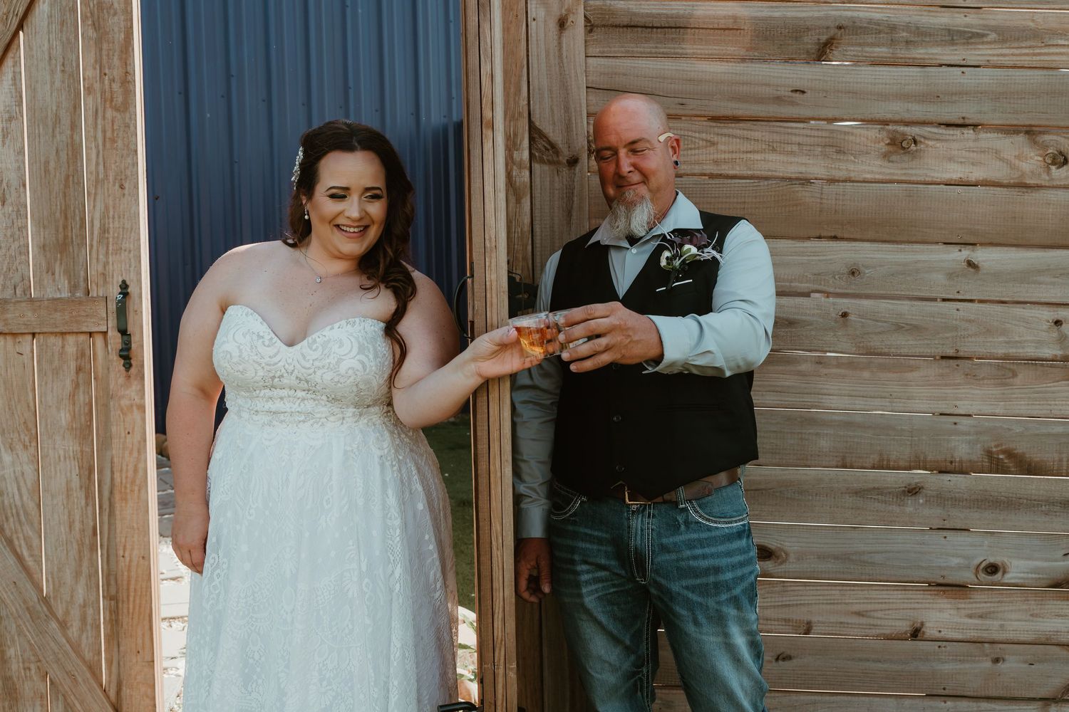 Bride in white gown and groom in vest toast with whiskey glasses outside a rustic barn.