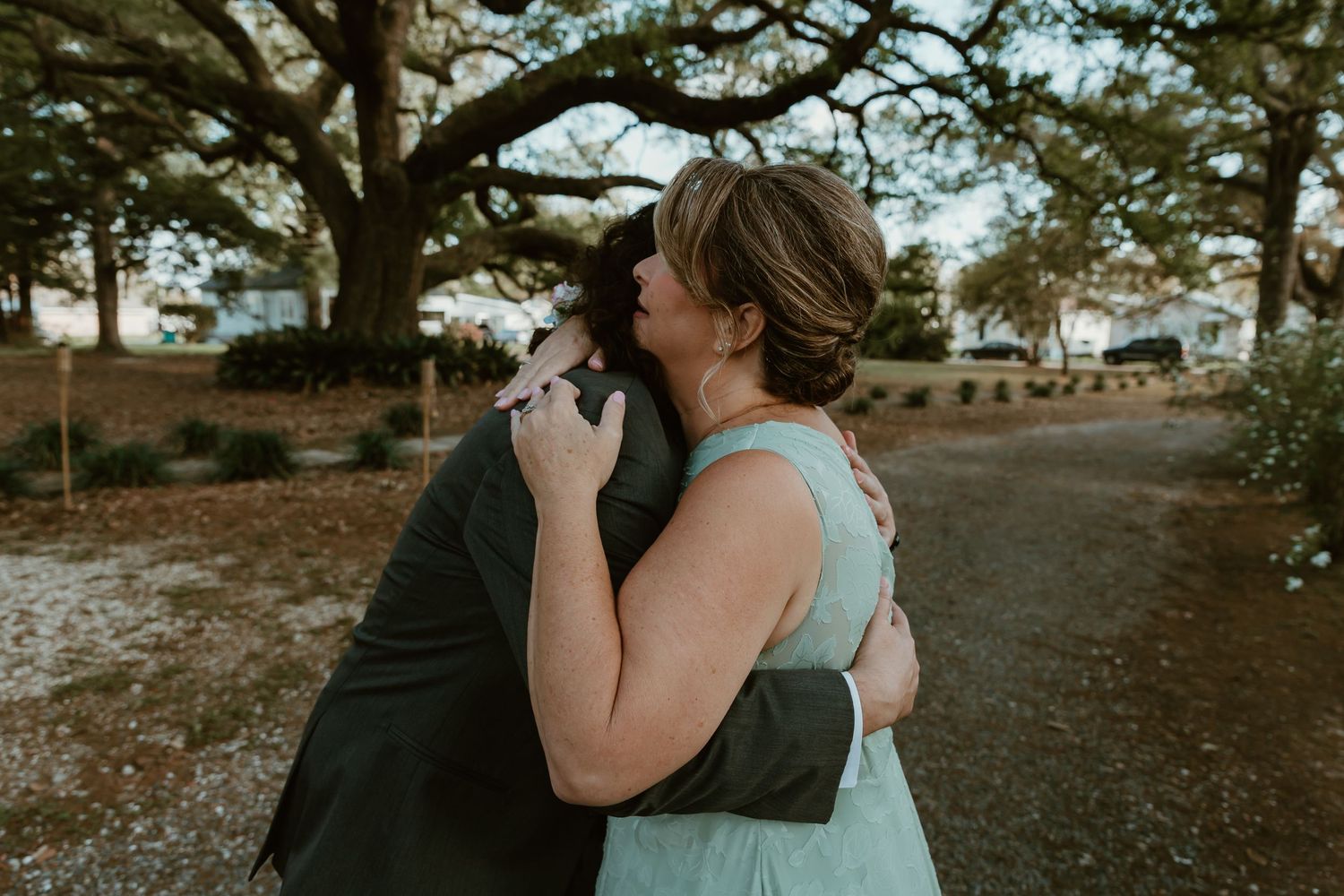Emotional outdoor embrace between mom and son in elegant attire beneath a large oak tree.