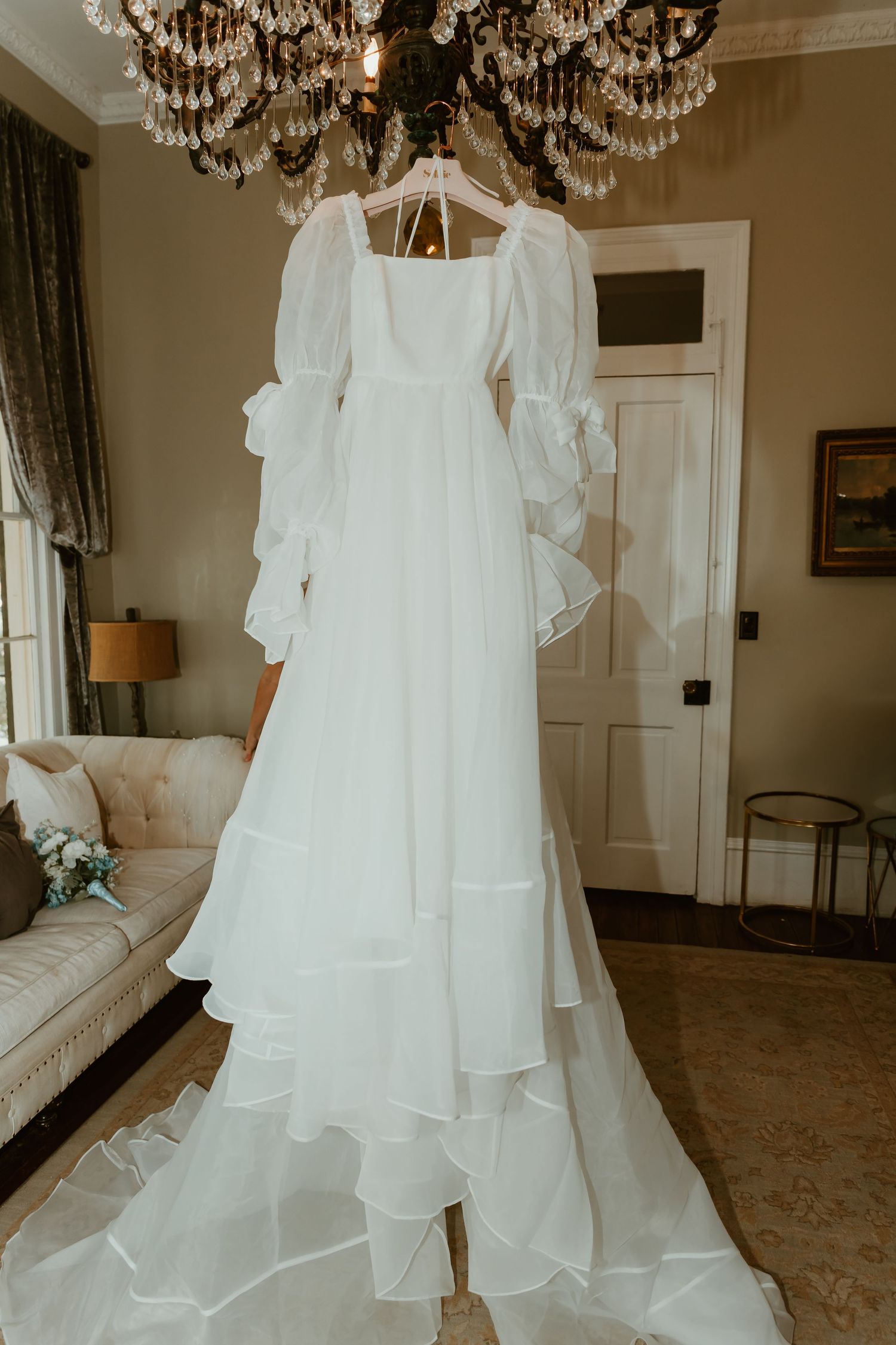 Elegant white bridal gown with voluminous sleeves and layered ruffle train displayed on crystal chandelier.