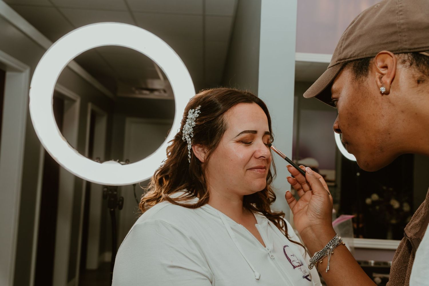 Bride gets professional makeup applied under a ring light while wearing a floral hair accessory.