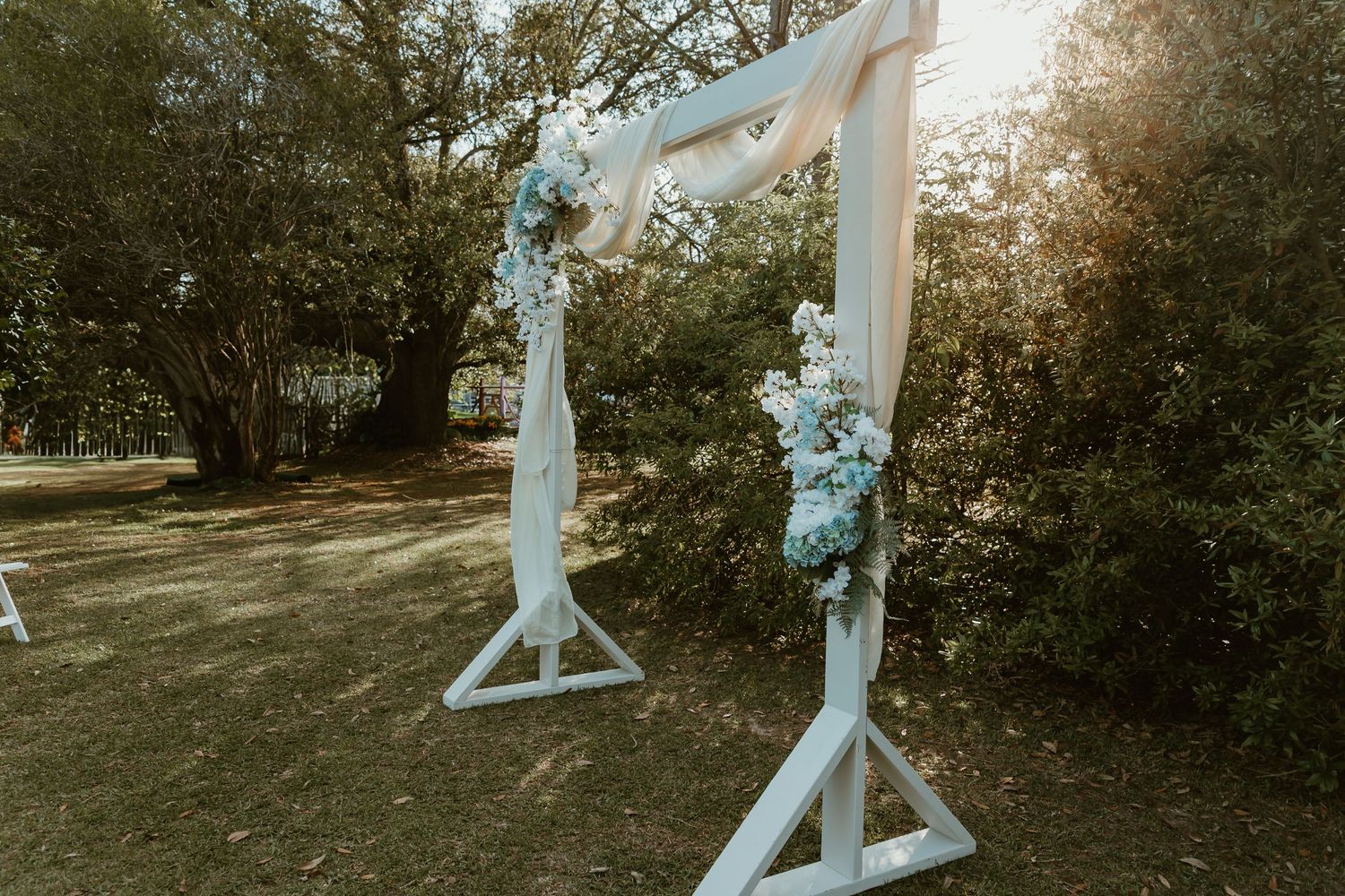 White wooden wedding arch decorated with blue and white flowers outdoors in dappled sunlight.
