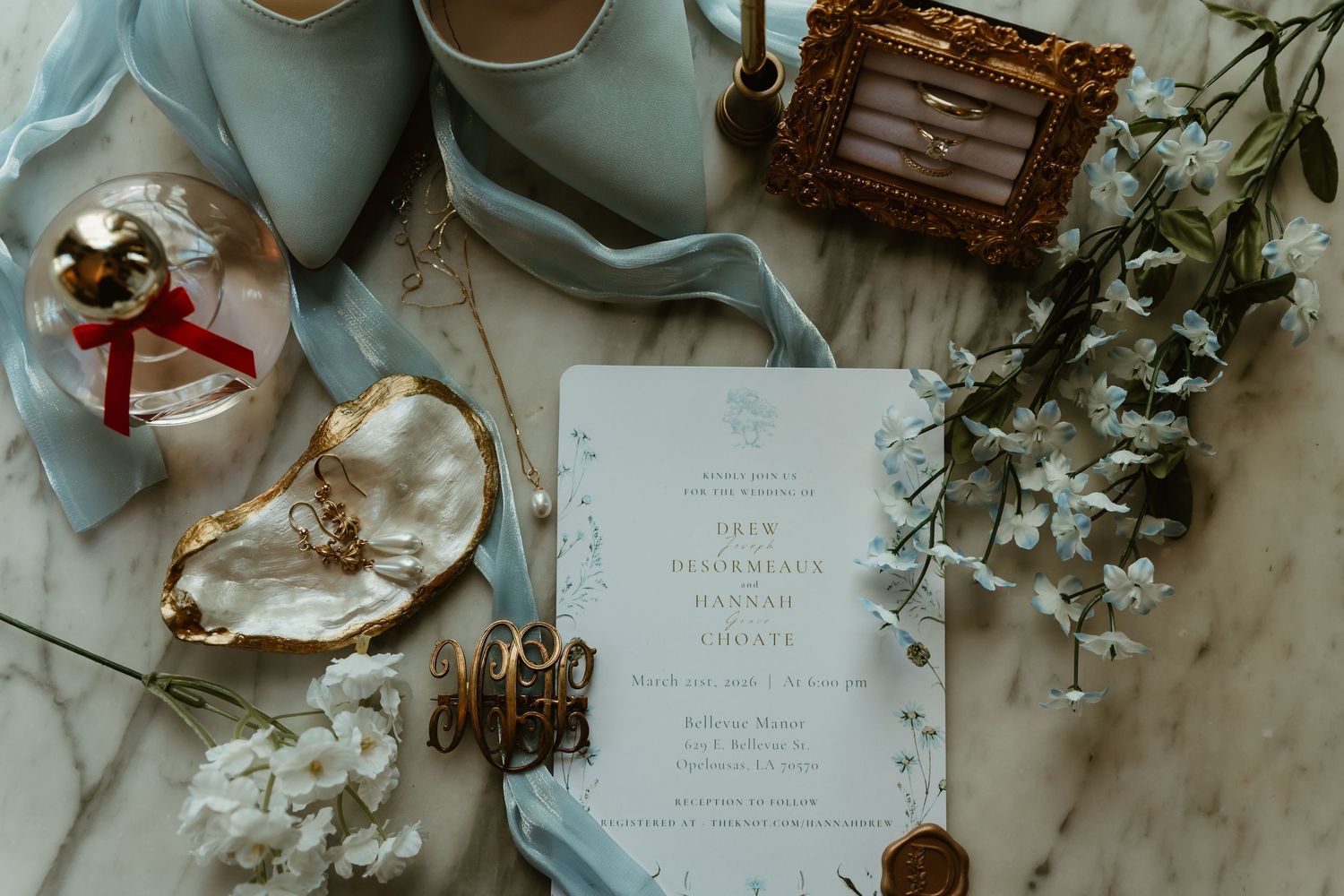 Elegant wedding flat lay with blue heels, invitation, rings, jewelry, and white florals on marble.