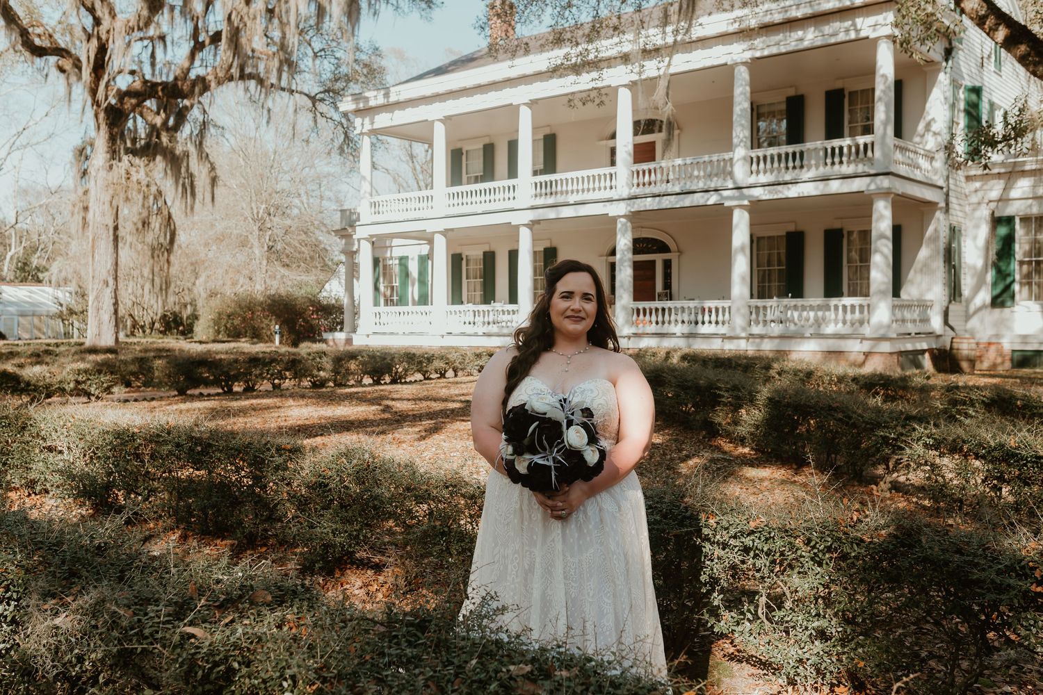Bride in strapless lace gown standing before a grand two-story Southern plantation home with manicured hedges.