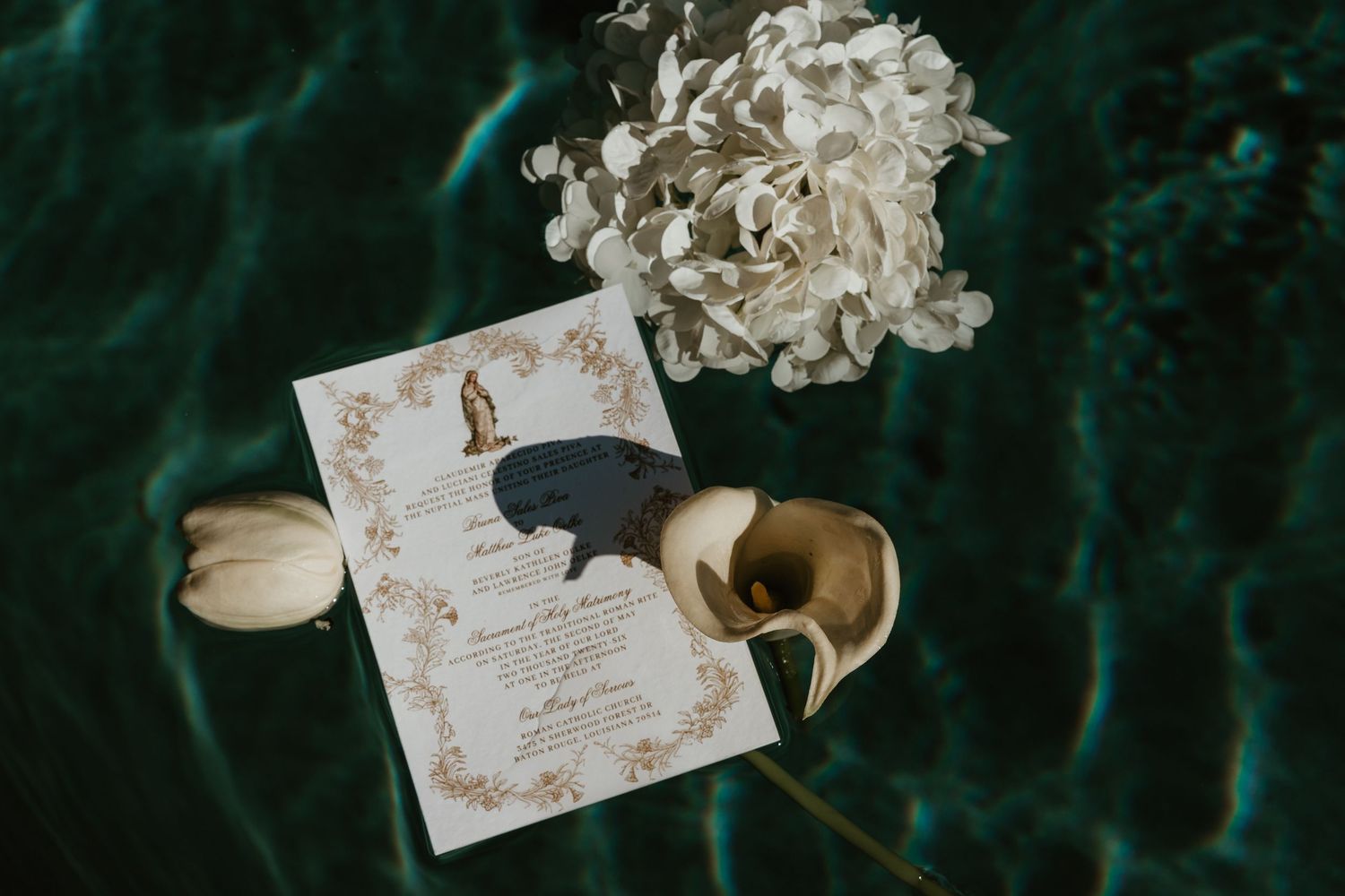 Wedding invitation with gold baroque border alongside white flowers in turquoise water.