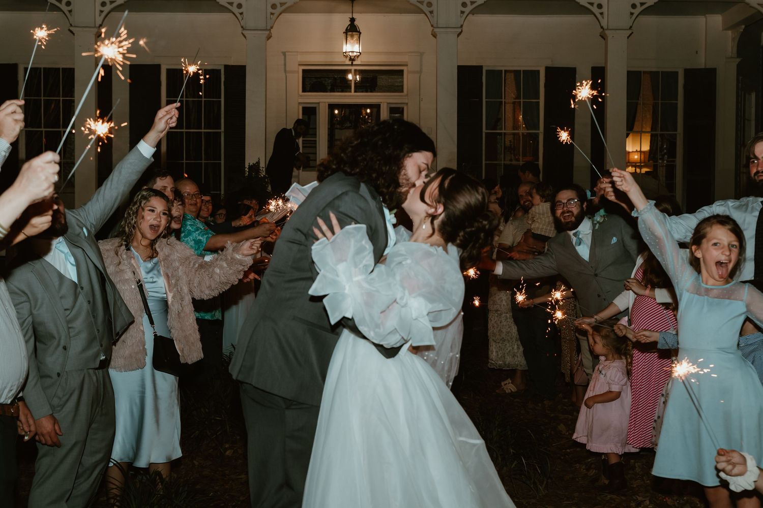 Bride and groom share romantic kiss during sparkler send-off surrounded by cheering guests at outdoor wedding reception.