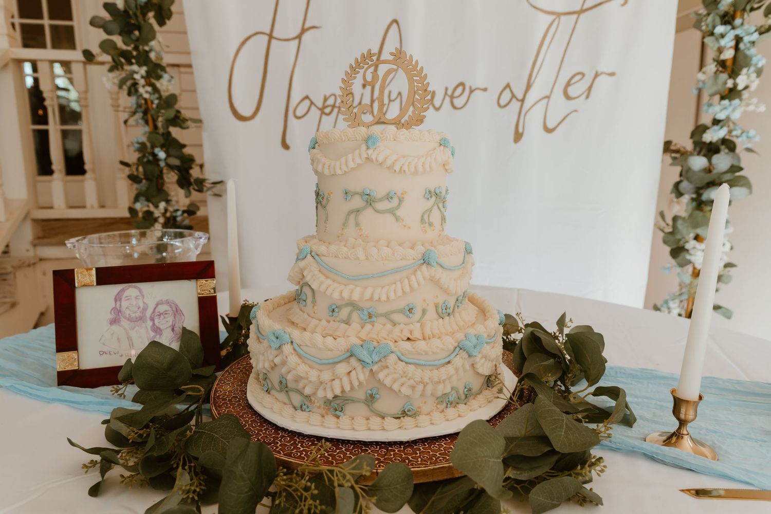 Three-tier white wedding cake with blue ribbon details and Happily Ever After topper on gold stand.