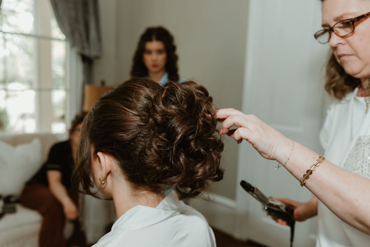 Hair stylist creating elegant updo hairstyle for bride during wedding preparation in bright room.