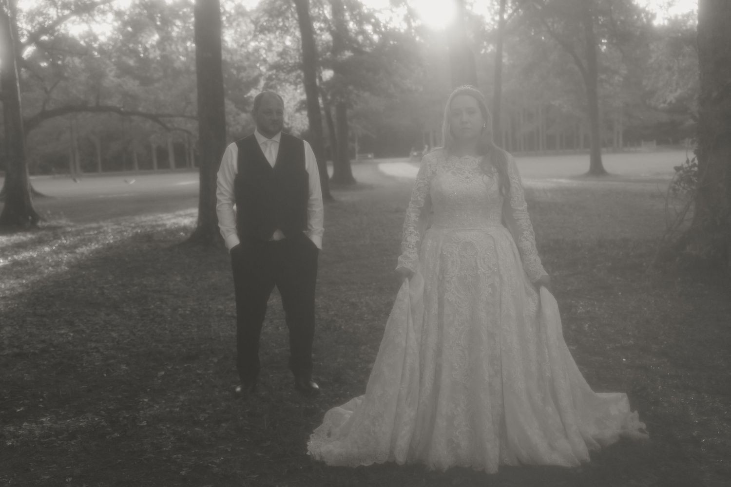 Black and white photo of bride in lace gown and groom in vest standing among trees at sunset wedding.