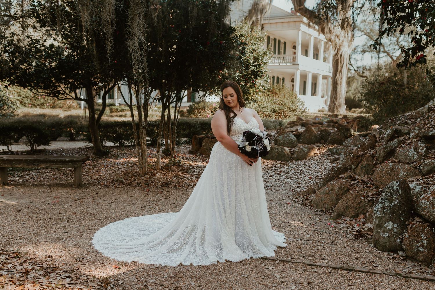 Bride in flowing white lace wedding dress with long train posing outdoors near historic mansion.