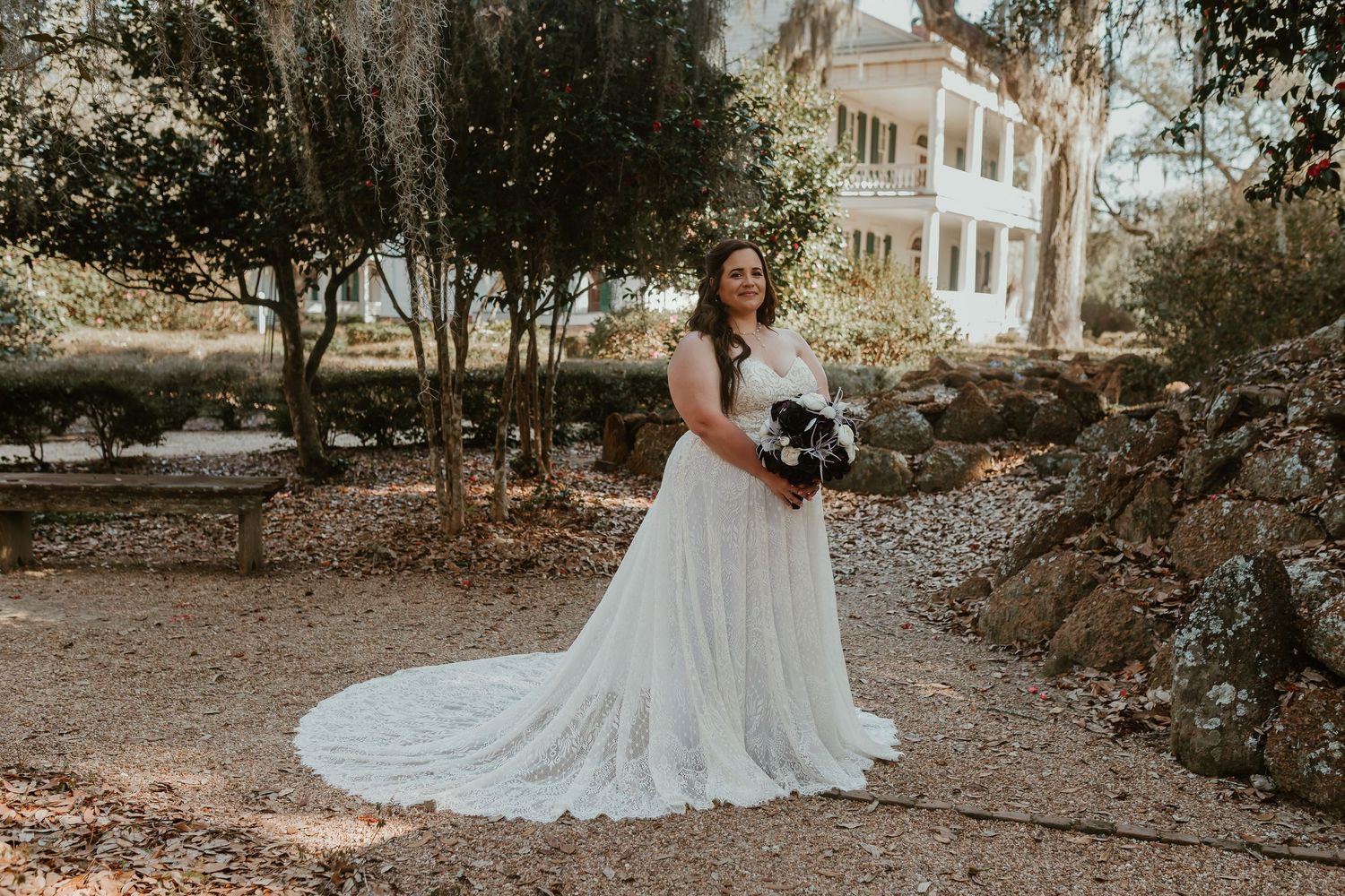 Bride in elegant white lace wedding dress with long train standing in sunlit garden near mansion.