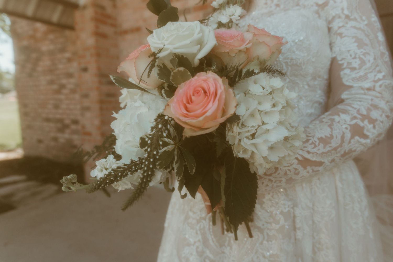 Bride holds a stunning wedding bouquet featuring pink roses and white hydrangeas in a lace gown.