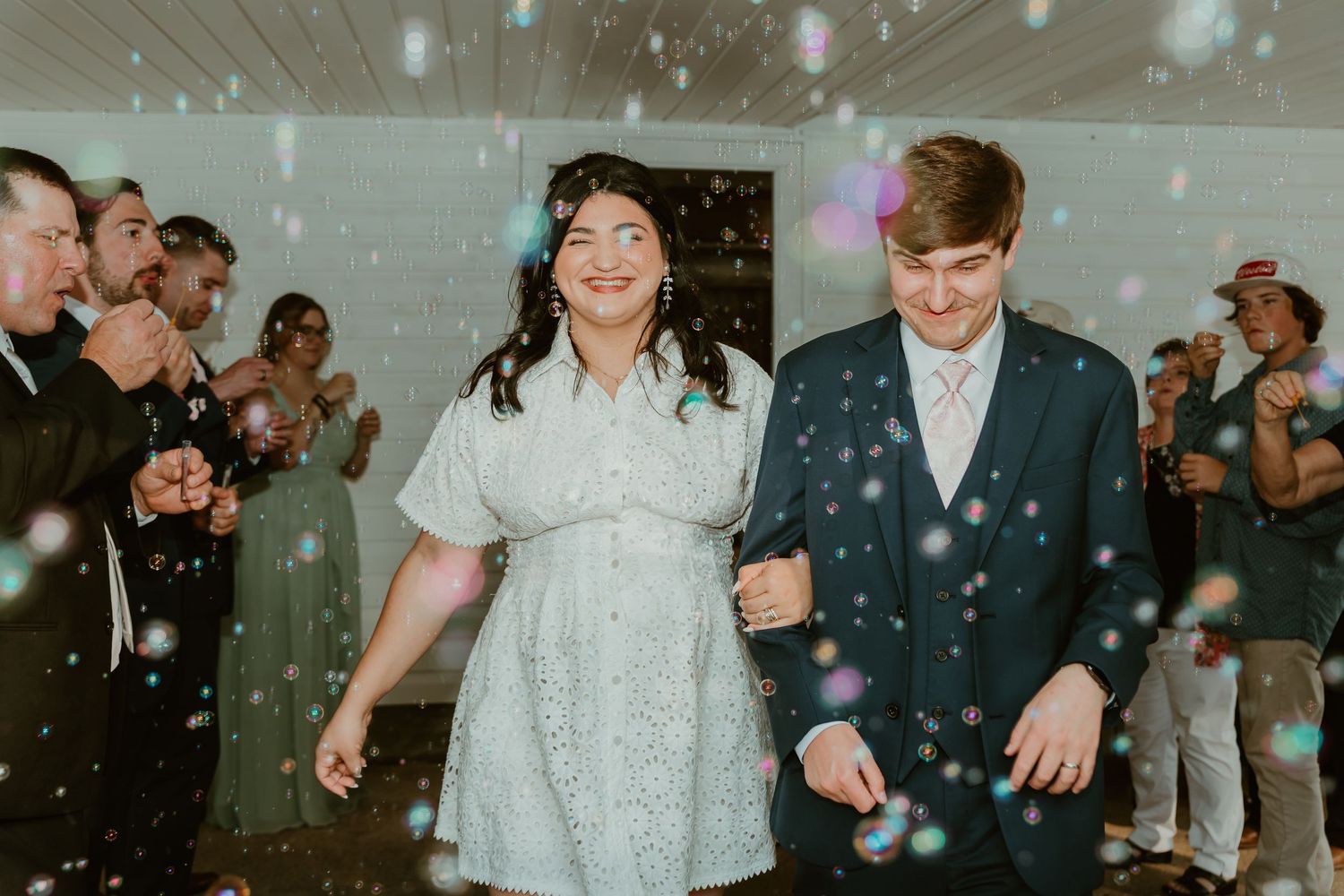Bride and groom smile while walking through bubbles during indoor wedding reception celebration.