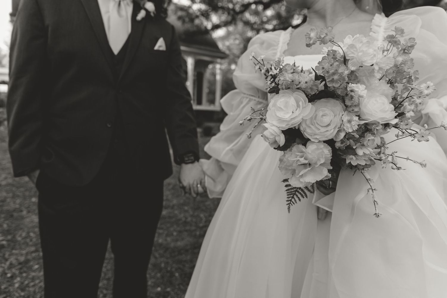 Black and white photo of a bride and groom holding hands, bride carries a large floral bouquet outdoors.