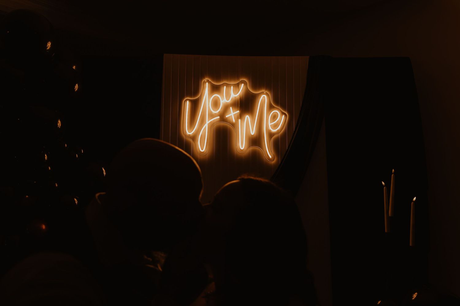 Couple silhouetted in front of a glowing orange You and Me neon sign in a dark room.