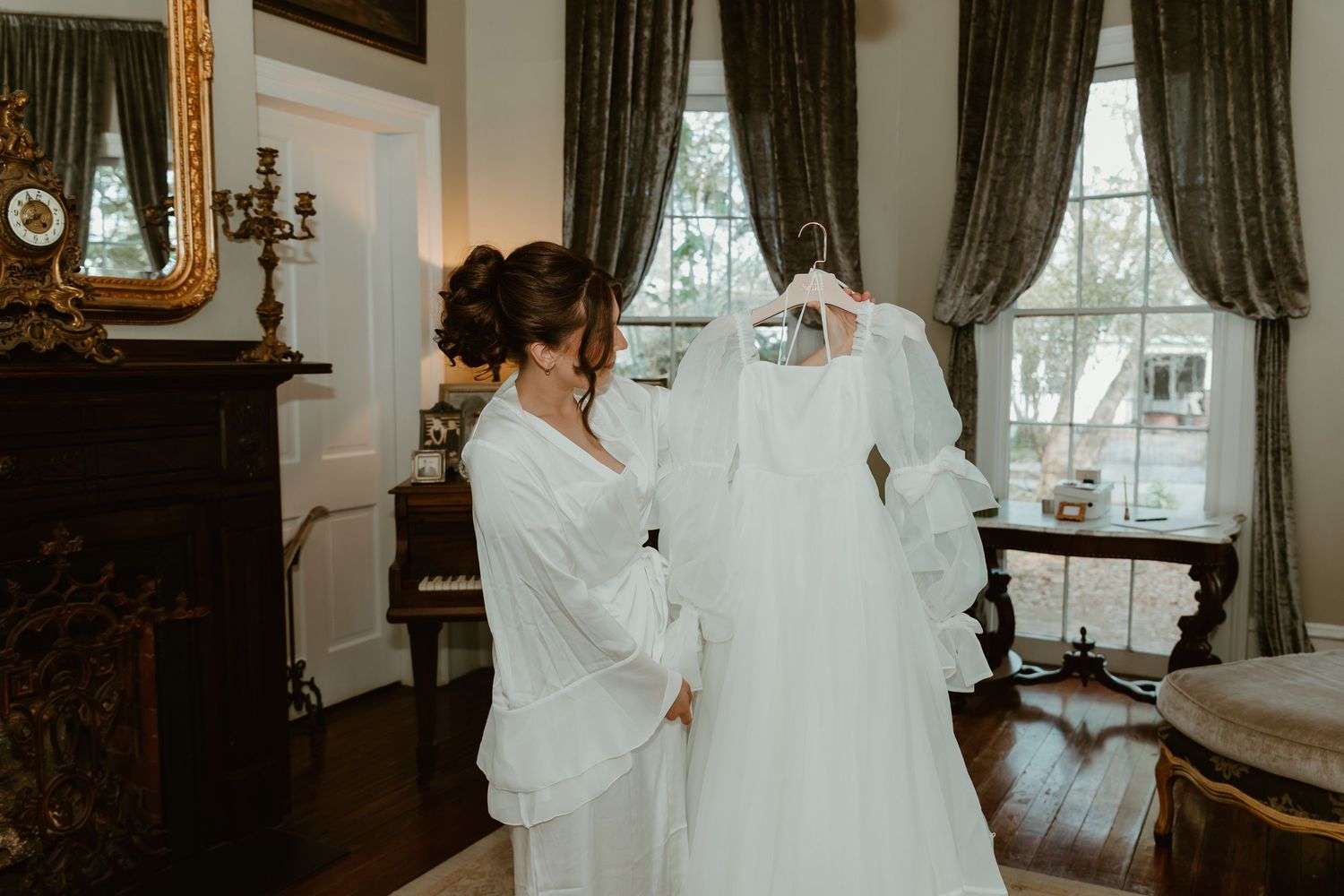 Bride in white robe admiring her wedding gown hanging near window in elegant room.