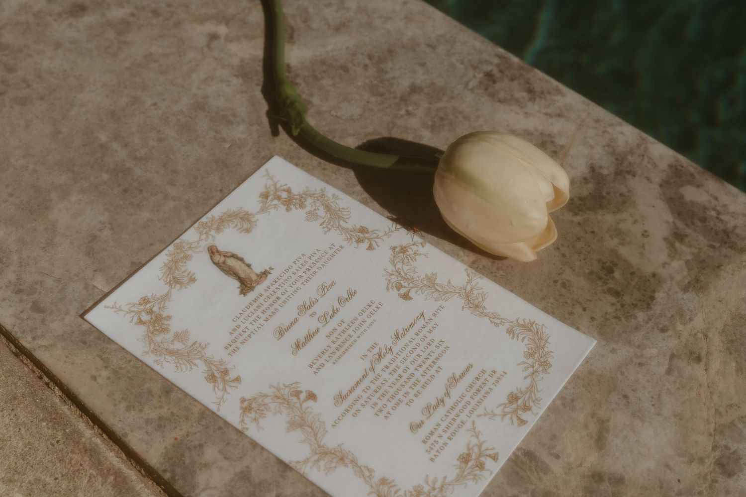 Wedding invitation with gold decorative border displayed on stone surface beside a white tulip.