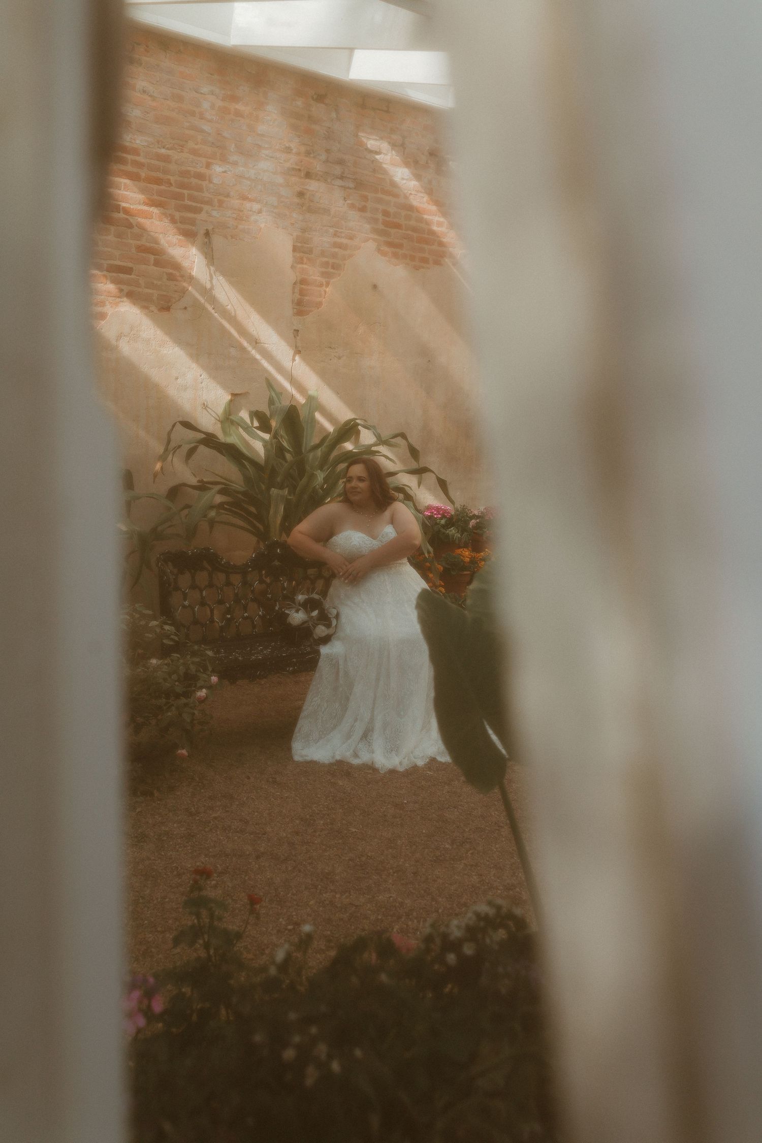 Bride in white lace wedding dress holding colorful bouquet, viewed through window in garden courtyard.
