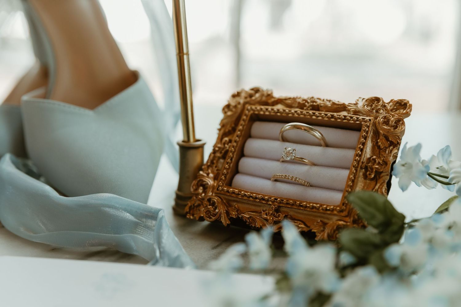 Gold ornate picture frame displaying wedding rings with blue floral details and greenery in background.
