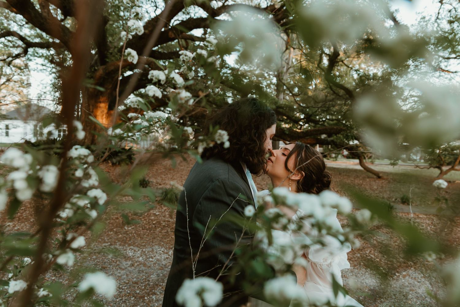 A bride and groom share a romantic kiss framed by blooming white flowers in a garden setting.